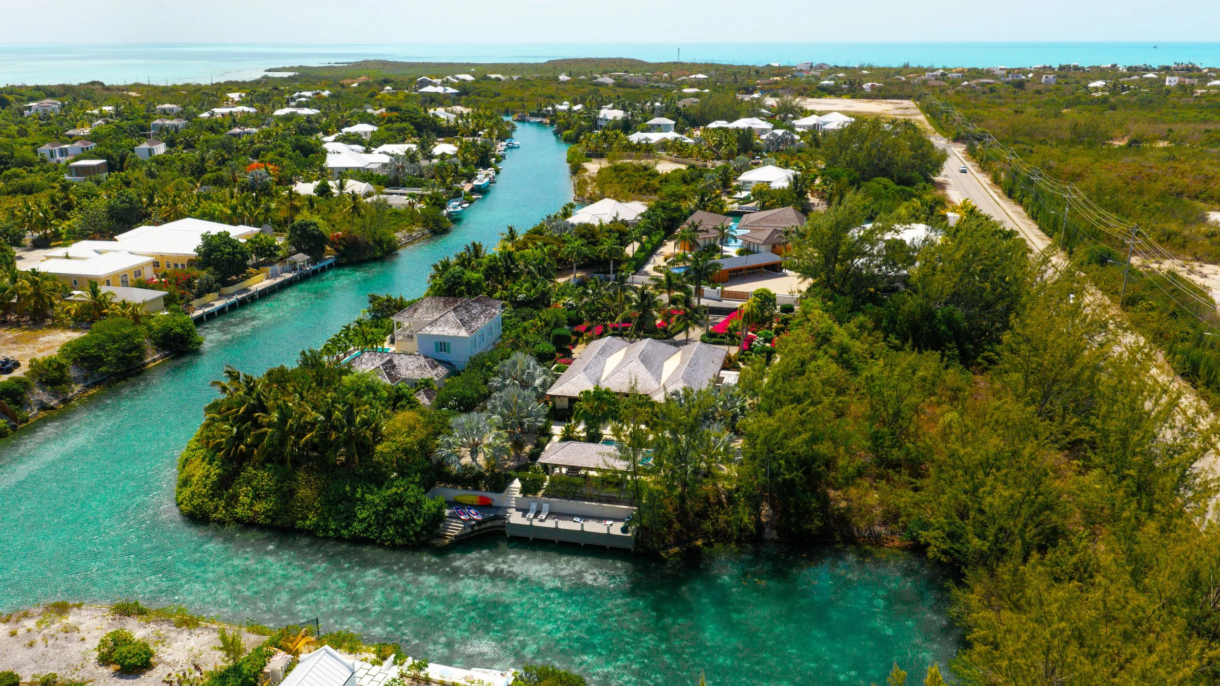 Aerial view of lush green tropical houses along a narrow turquoise water canal near the coast, with some boats docked and trees surrounding the area.