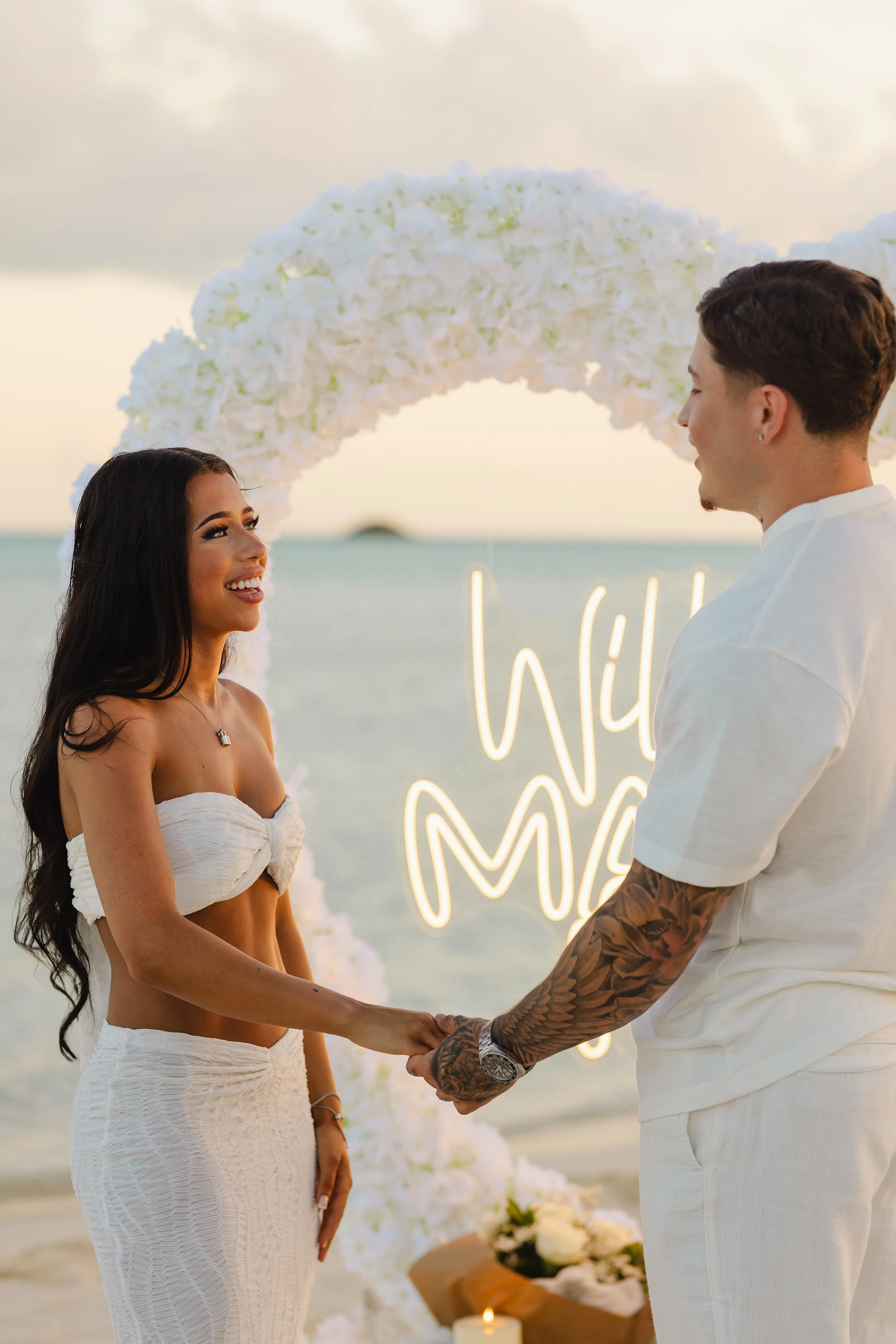 A couple holding hands and exchanging vows during a beach wedding, with an arranged floral arch and beach setting in the background.