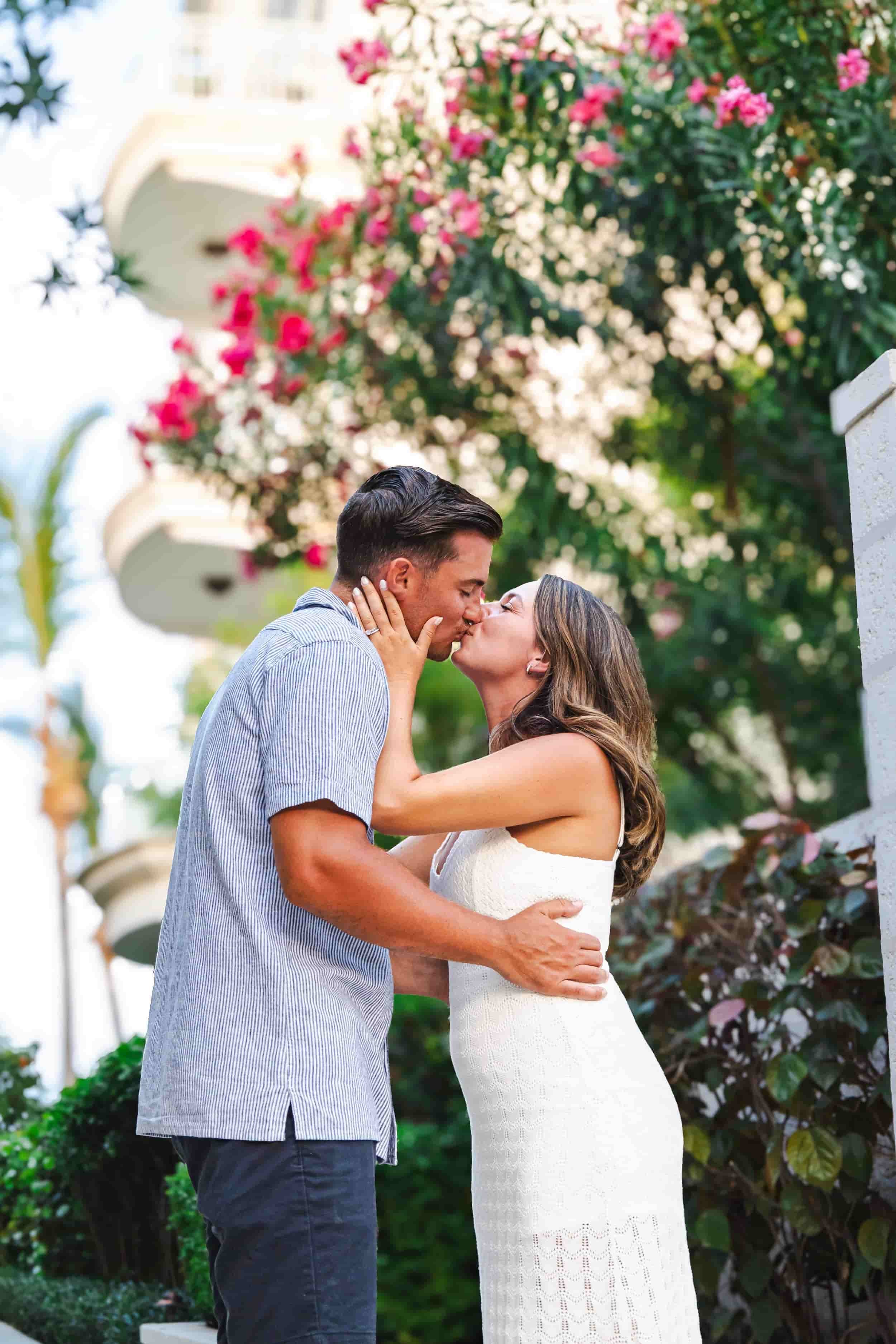 A couple sharing a kiss outdoors with blooming pink and white flowers in the background.