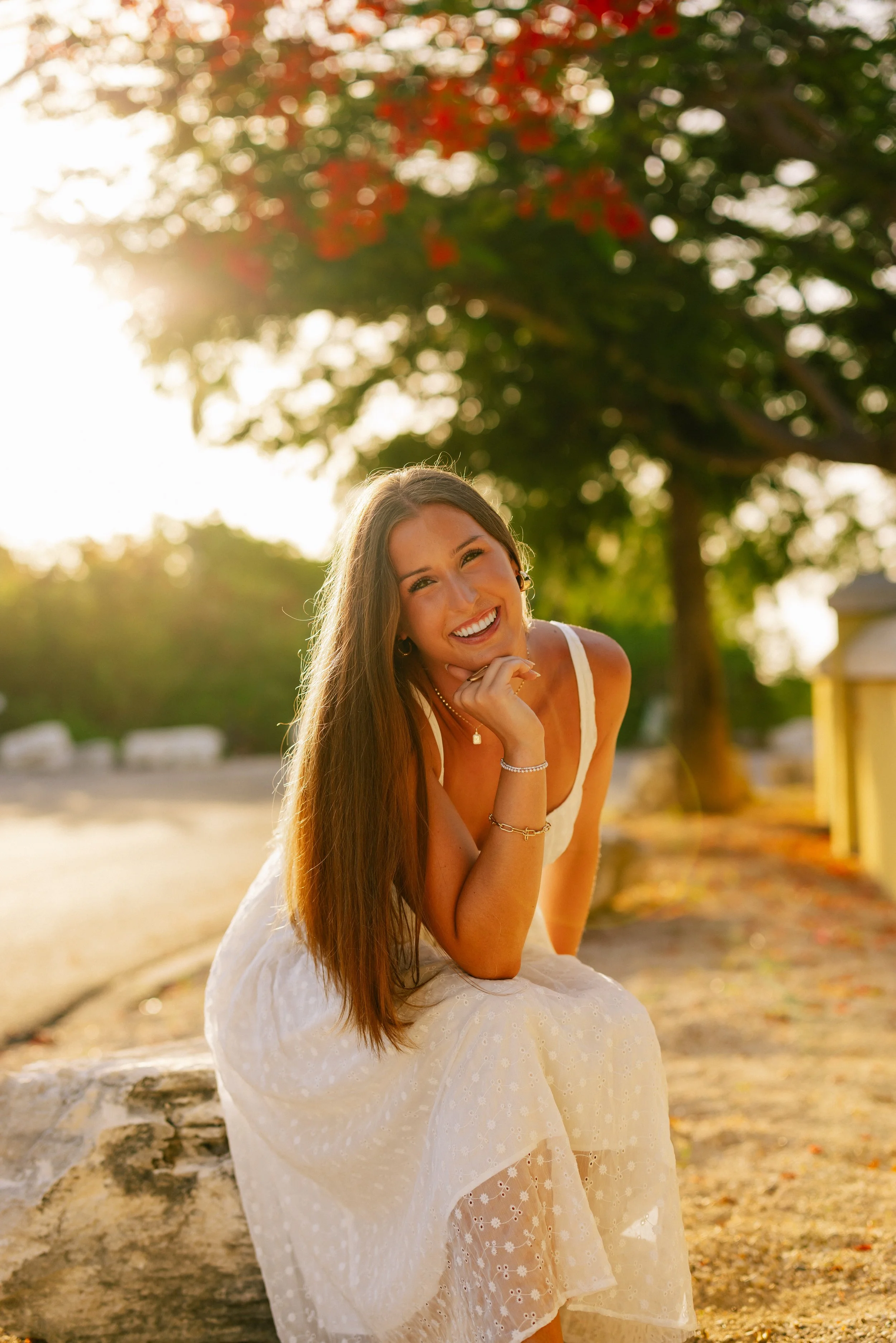 A young woman with long brown hair smiling and sitting outdoors on a sunny day, wearing a white dress with eyelet fabric, surrounded by green trees with sunlight filtering through their leaves.
