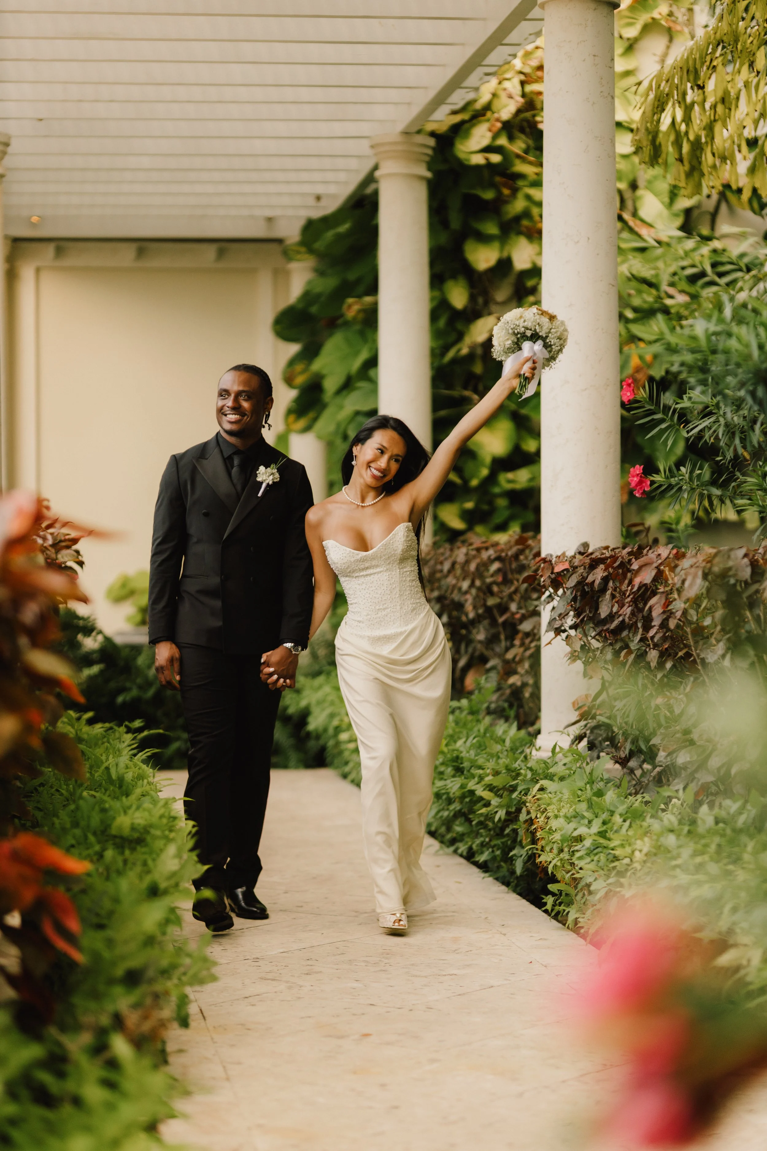 A newlywed couple walking hand in hand on a garden pathway surrounded by lush green plants and colorful flowers, with the bride holding a bouquet in the air and smiling.