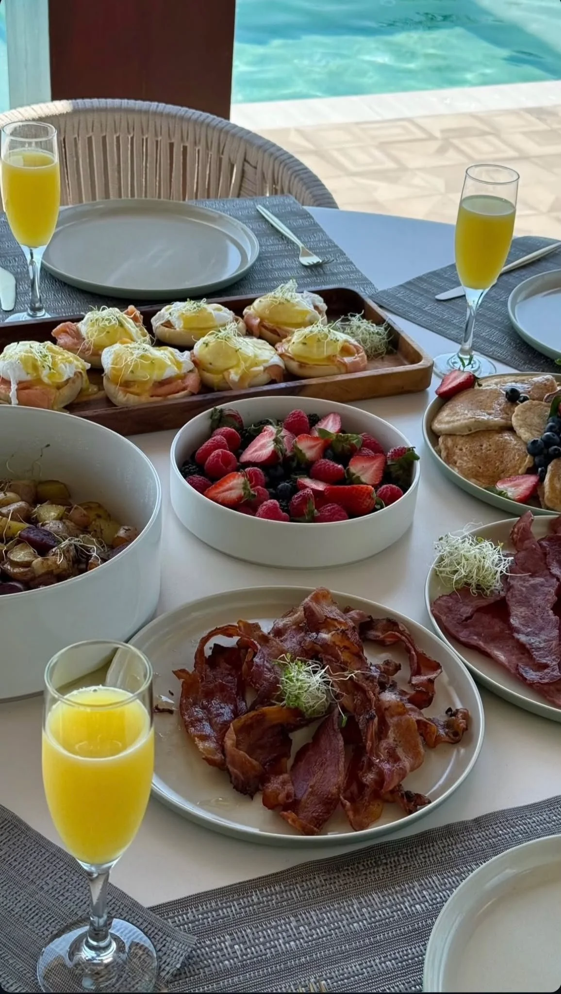 A breakfast table with various foods including scrambled eggs with prosciutto and sprouts, fresh berries, sliced meats, crispy bacon, and pancakes. There are two glasses of orange juice and some empty plates with utensils near a pool.