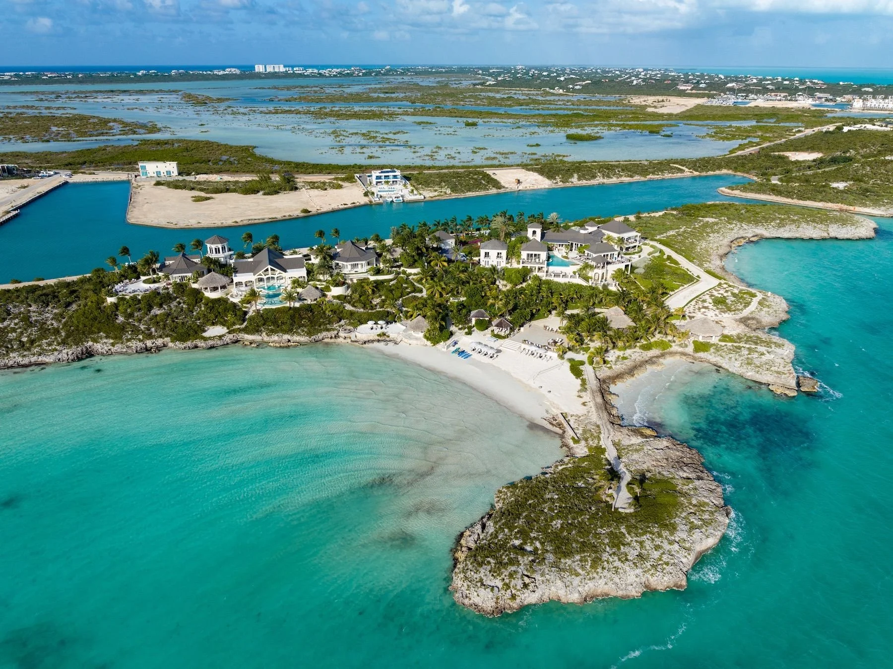 Aerial view of a tropical coastal area featuring a sandy beach, turquoise waters, lush greenery, luxury houses, and waterways with some boats at the edge of the shoreline.