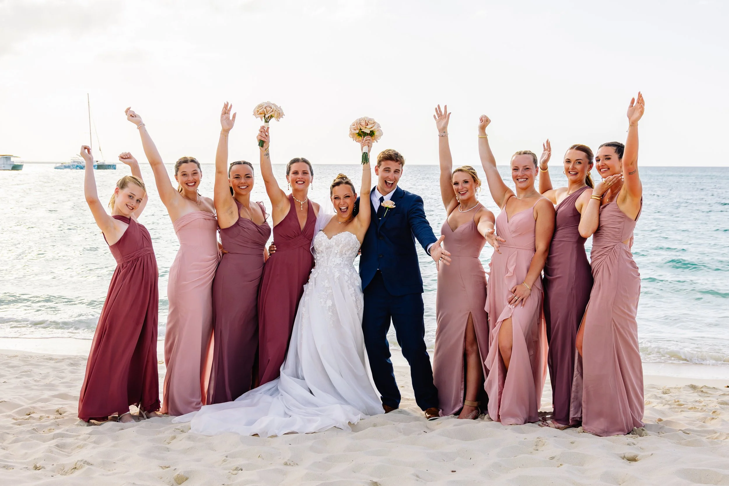 A bride and groom with their wedding party on a beach, all smiling and holding bouquets, celebrating the wedding.