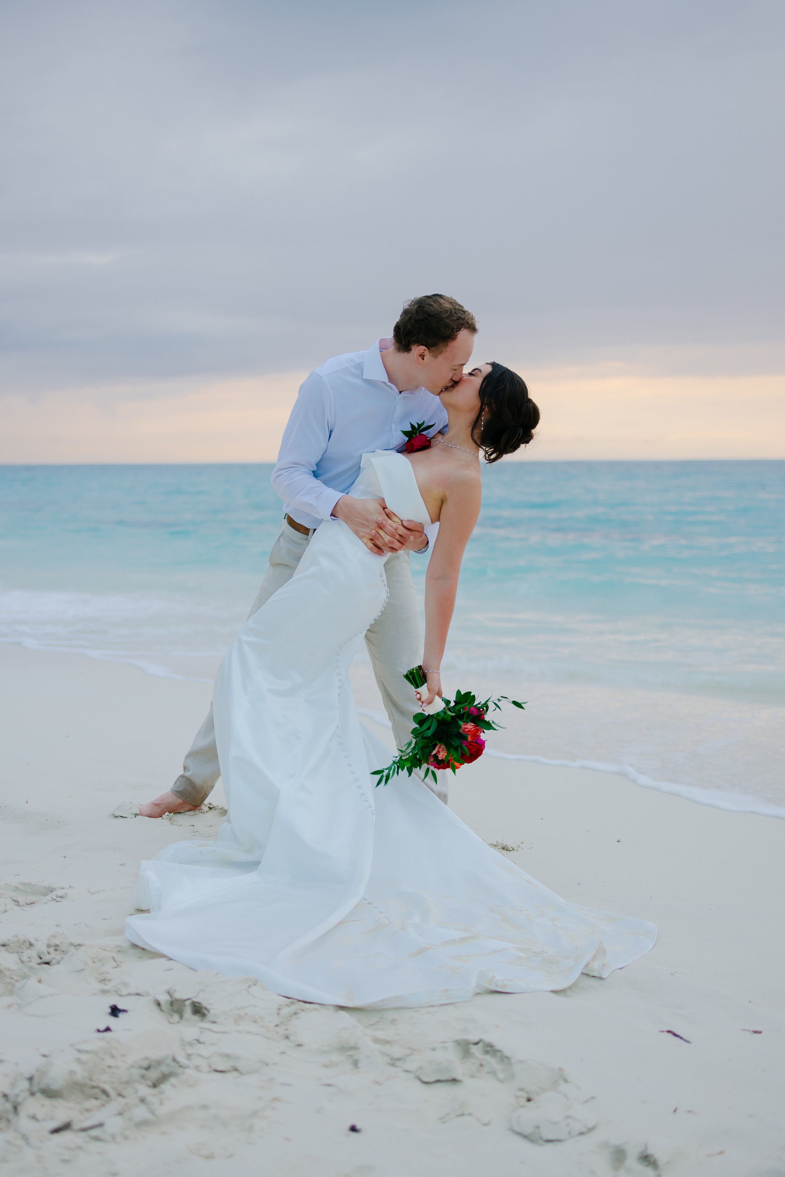 Couple in wedding attire sharing a kiss on a beach with ocean waves and cloudy sky in the background.