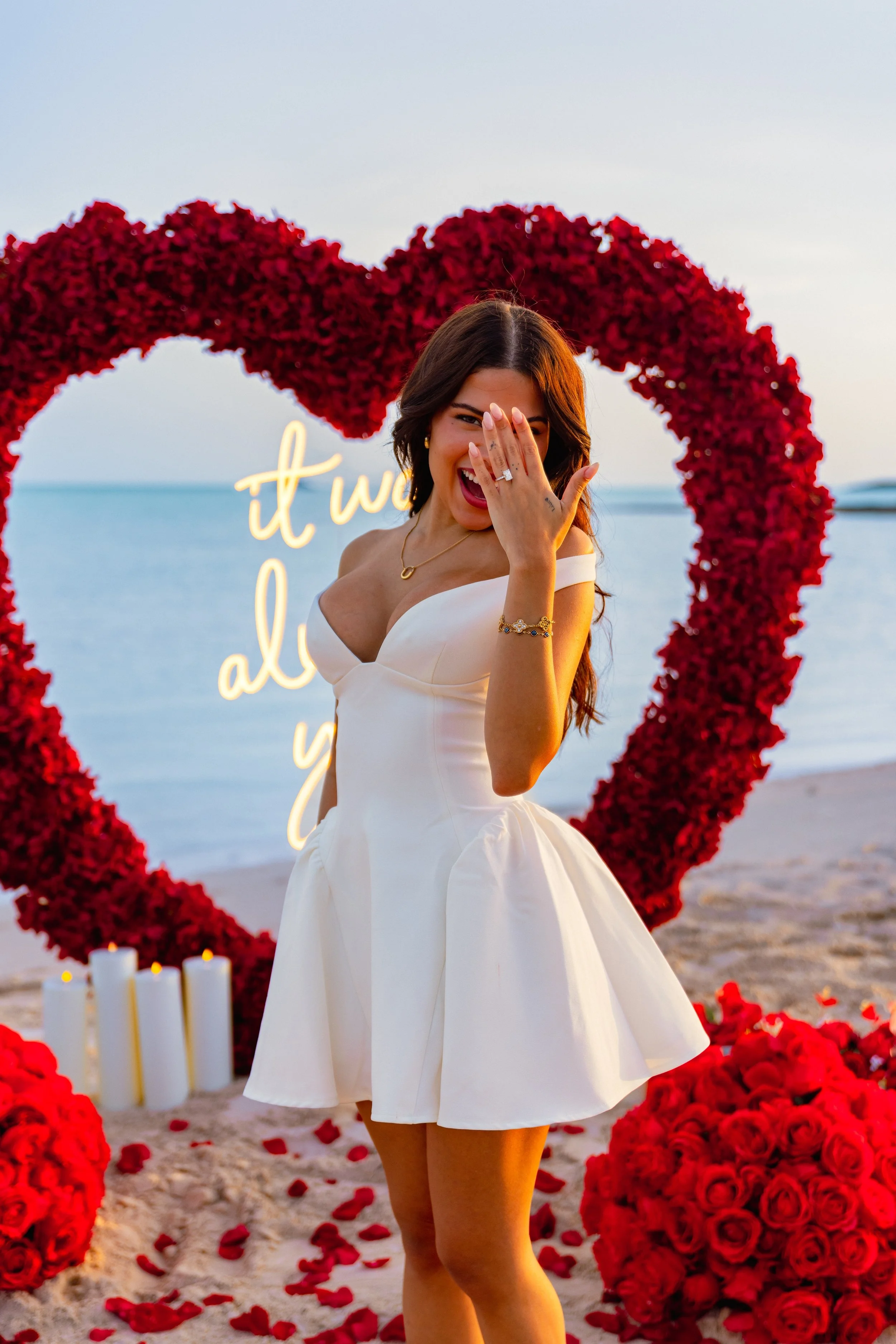 A woman in a white dress smiles and covers her mouth with her hand at a beachside wedding with a heart-shaped rose decoration and candles in the background.