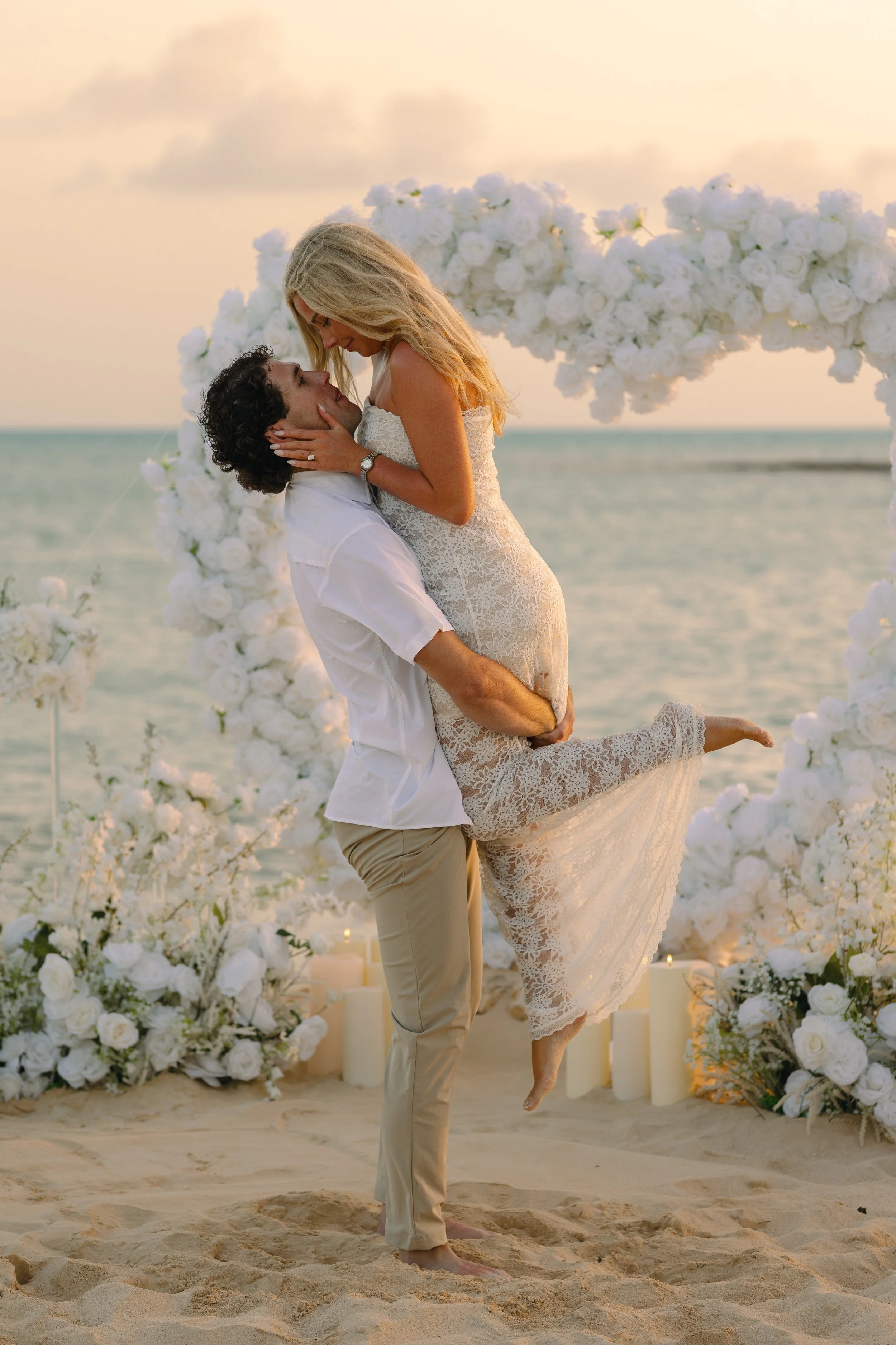 A couple at a beach wedding, with the man lifting the woman, who is wearing a white lace dress and the man in beige pants and a white shirt, in front of a floral arch made of white roses, candles, and greenery, during sunset.