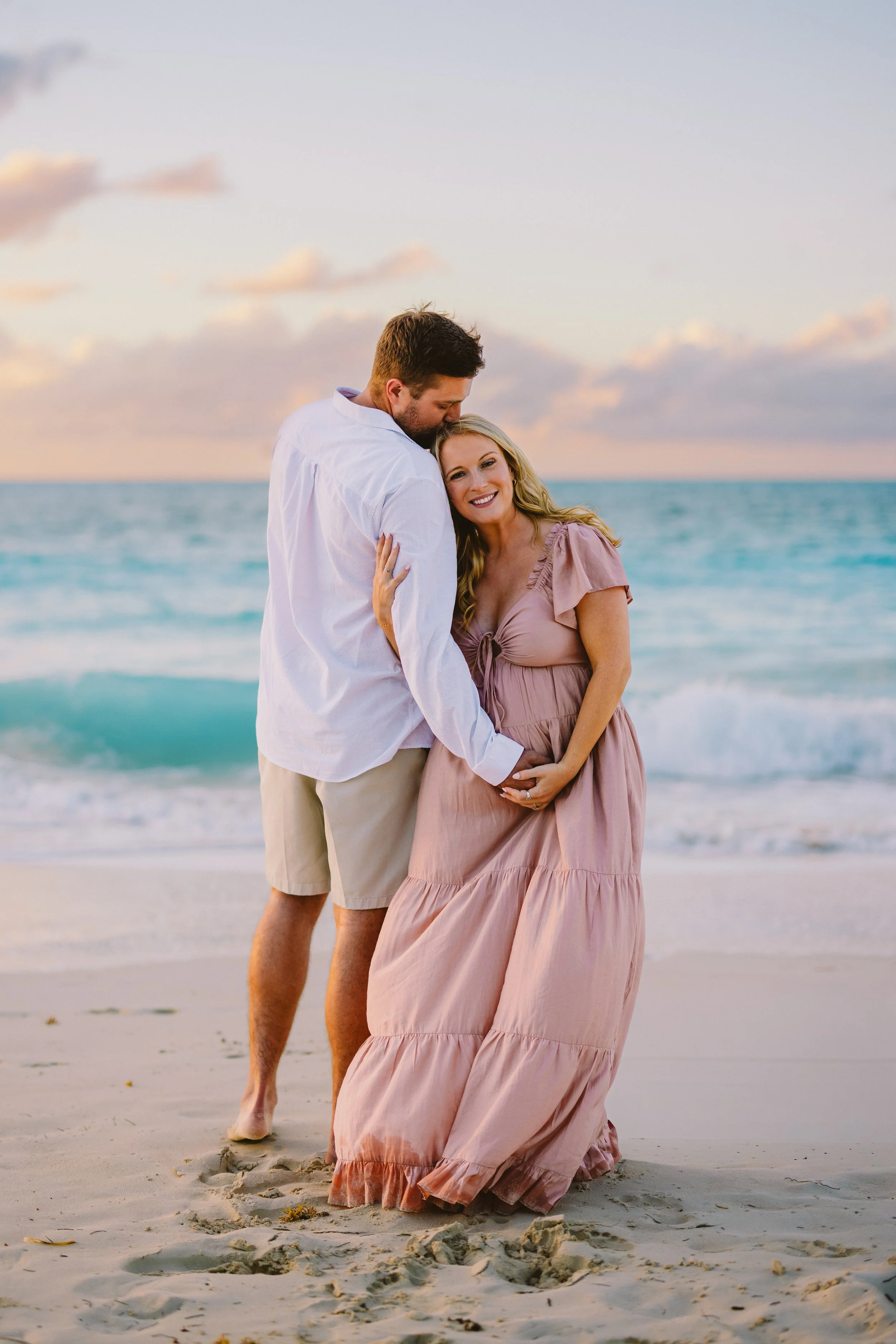 A couple stands on a sandy beach at sunset, with the man kissing the woman's head and she smiling while holding her pregnant belly.