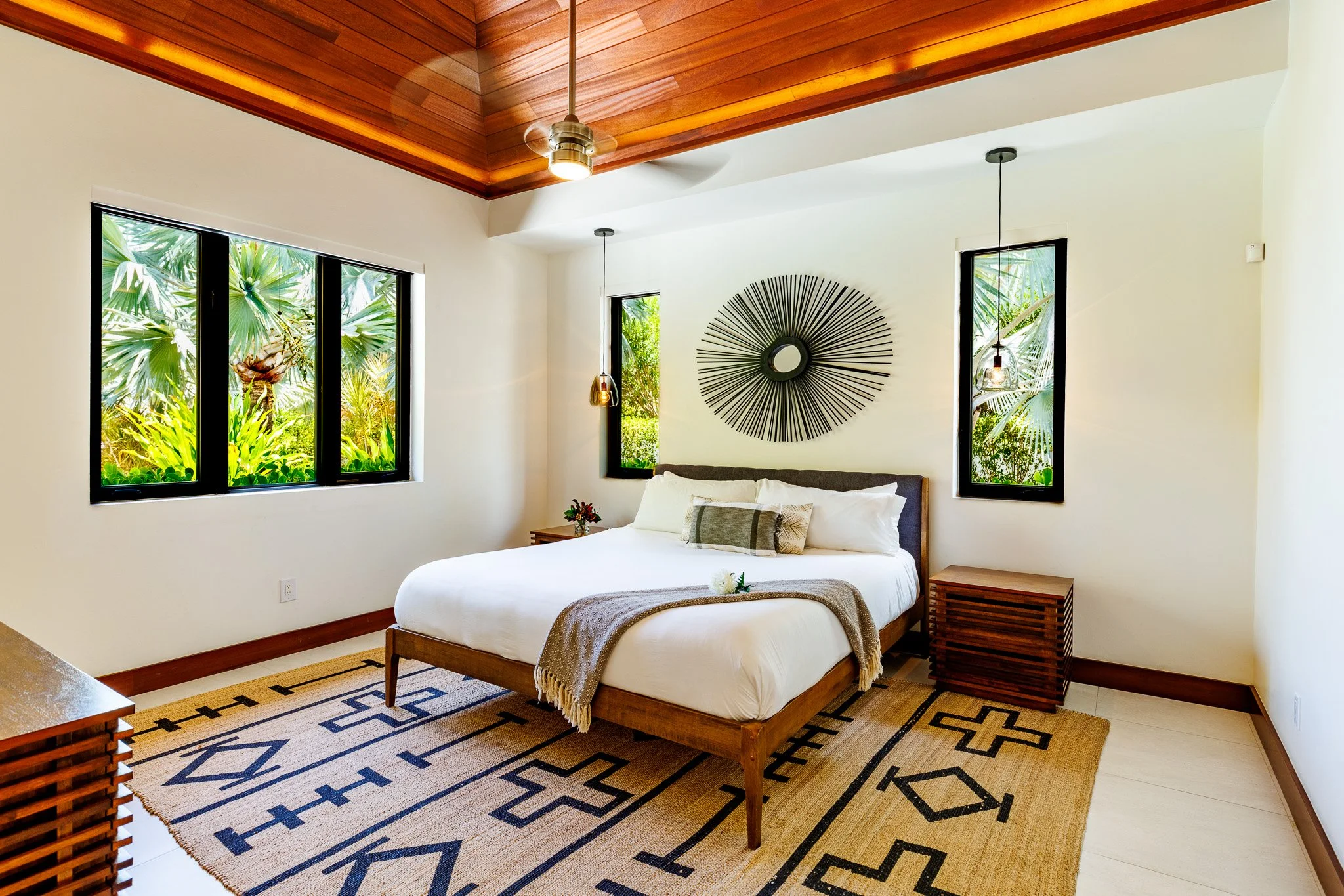 A bedroom with white walls, a wooden bed frame with a white mattress, a brown blanket, and a decorative pillow. There is a large wall art piece on the wall behind the bed. The room has three windows with views of green tropical plants outside. The ceiling is wooden with a modern ceiling fan and pendant lights hanging over the side tables. The floor has a beige and black geometric patterned rug.