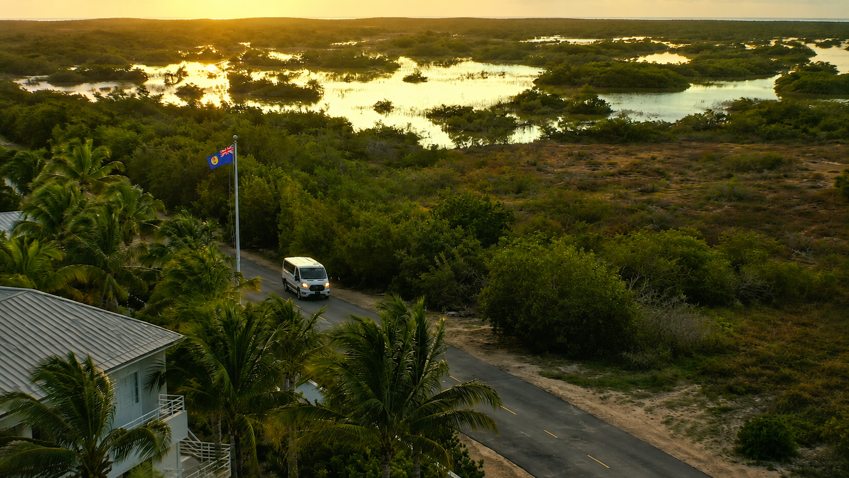 A white van driving along a narrow road lined with tall palm trees and lush greenery, near a flagpole flying the Australian flag, with a landscape of water and wetlands in the background during sunset.