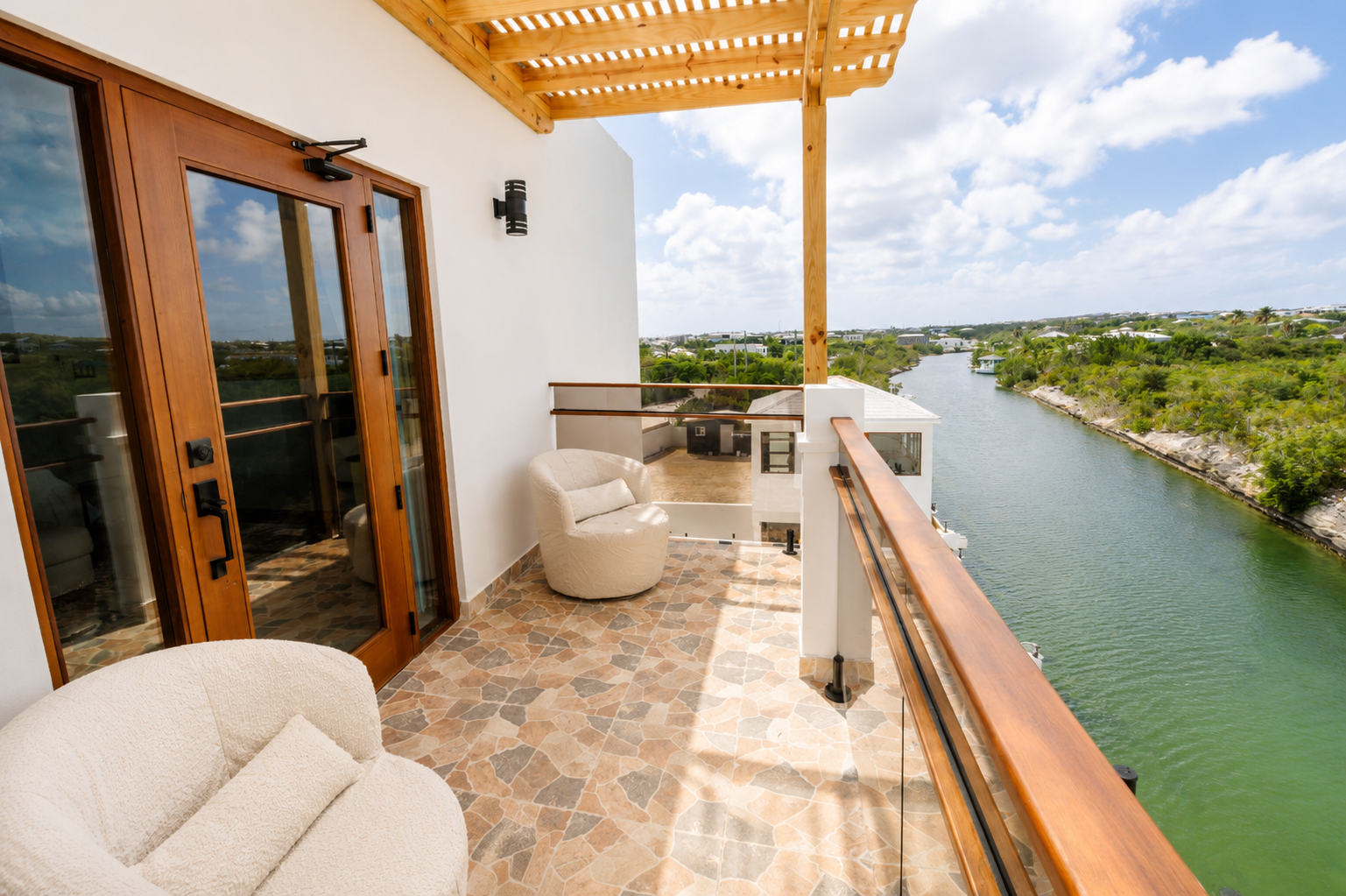 Balcony with wooden sliding doors, beige armchair, and railing overlooking a river and greenery on a partly cloudy day.