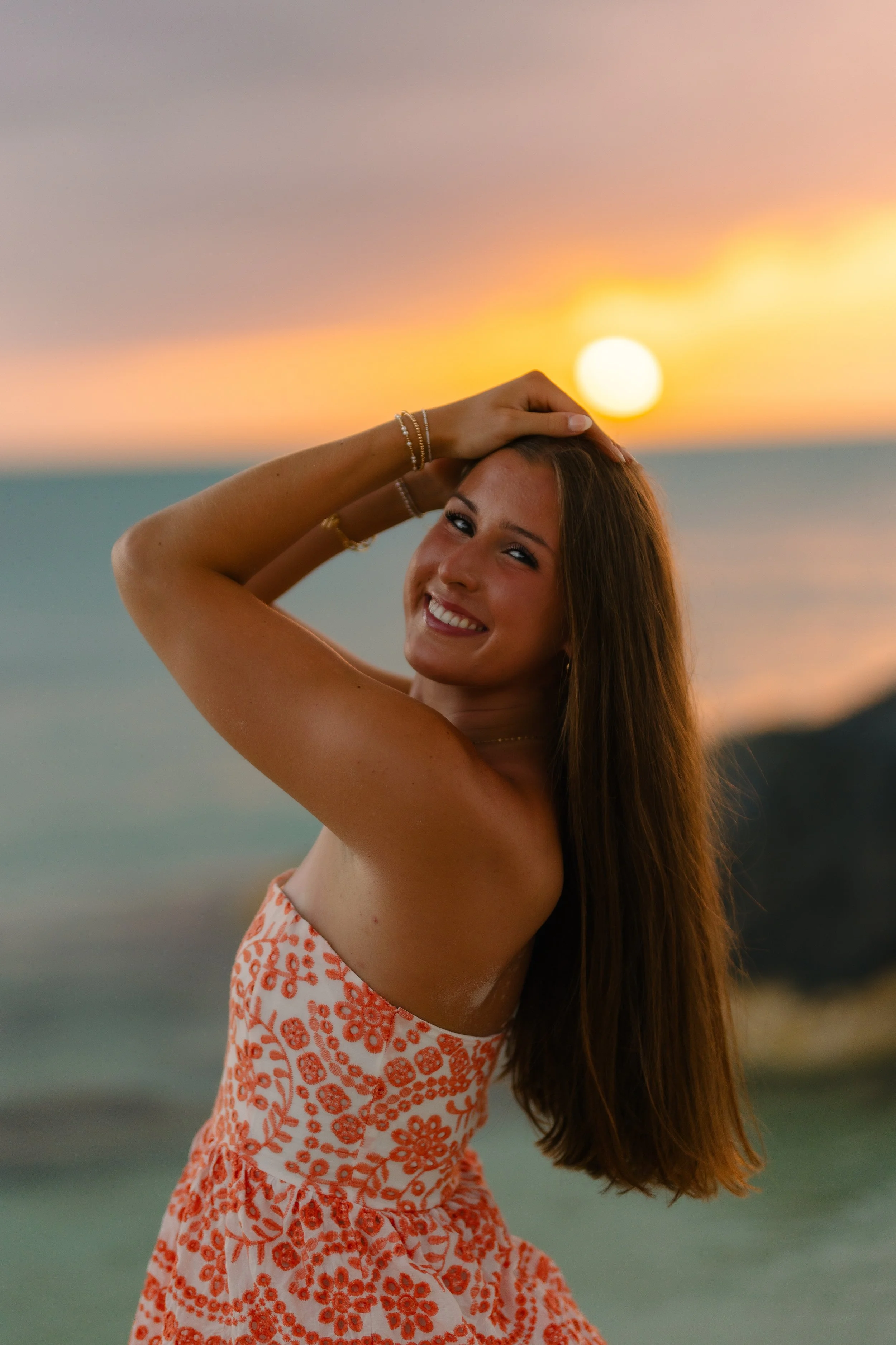 A young woman smiling at sunset at the beach, wearing a white and orange patterned dress, with her hand on her head, and long brown hair.