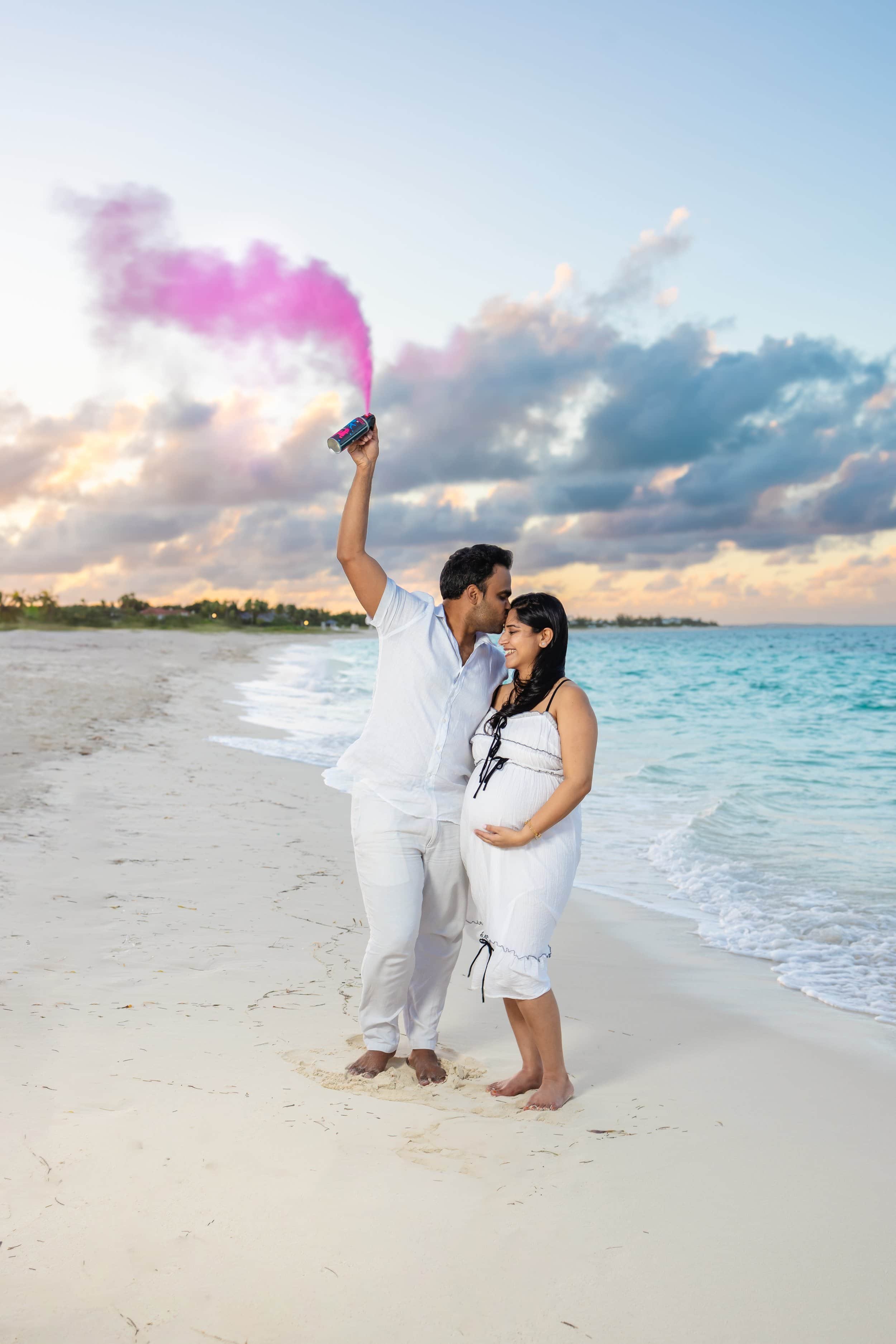 A pregnant woman and her partner standing on a beach, with the man holding a pink smoke bomb in the air, smiling and leaning in for a kiss.