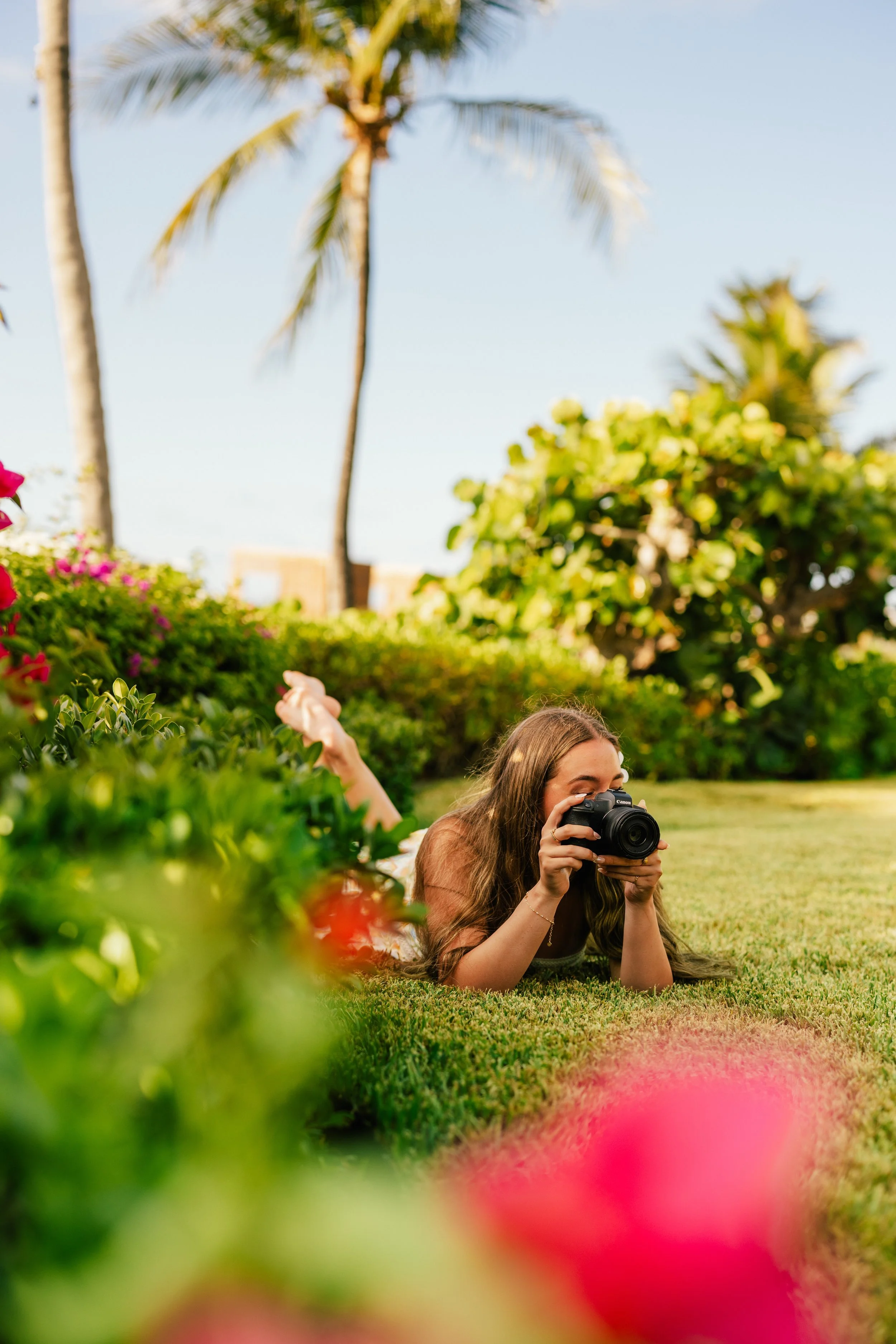 A woman with long hair lying on the grass, taking a photograph with a camera surrounded by colorful flowers and greenery, palm trees in the background under a clear sky.