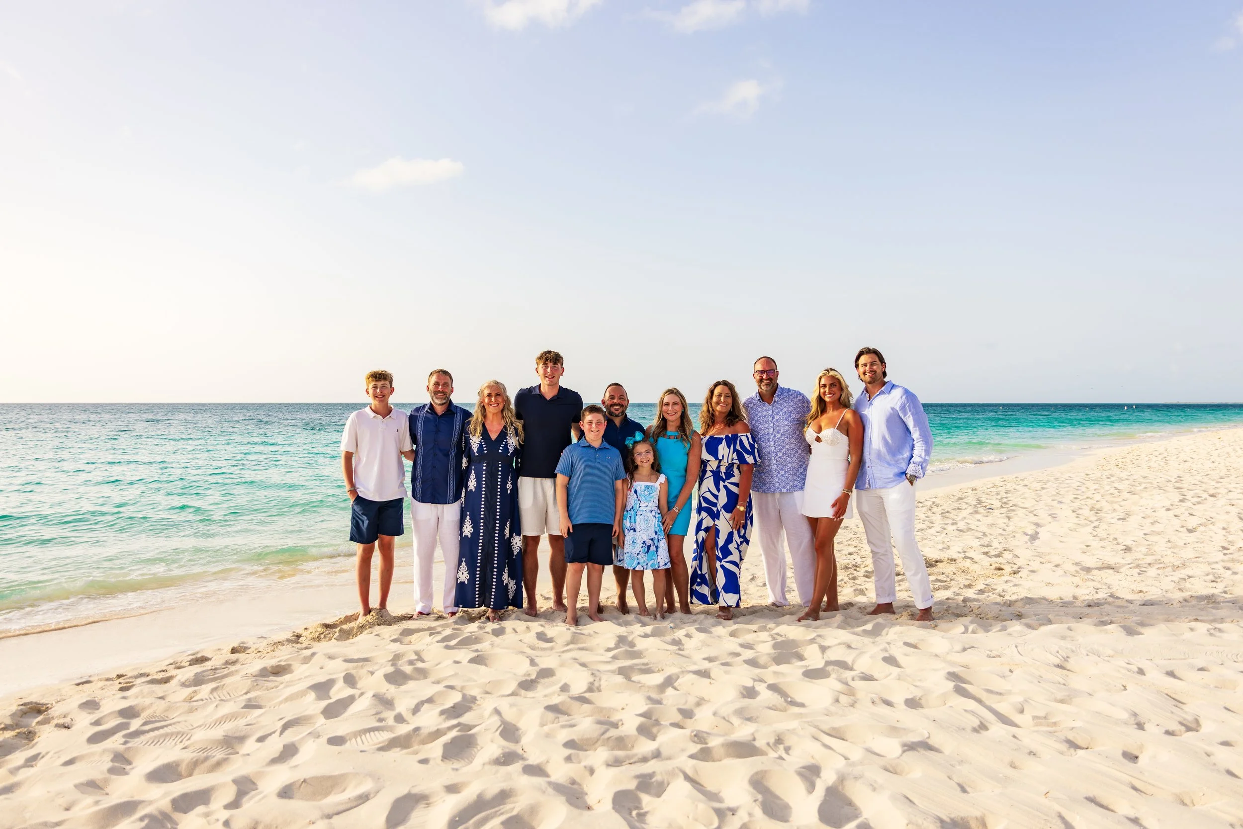 A large group of people standing on a sandy beach with the ocean in the background, smiling for a photo.