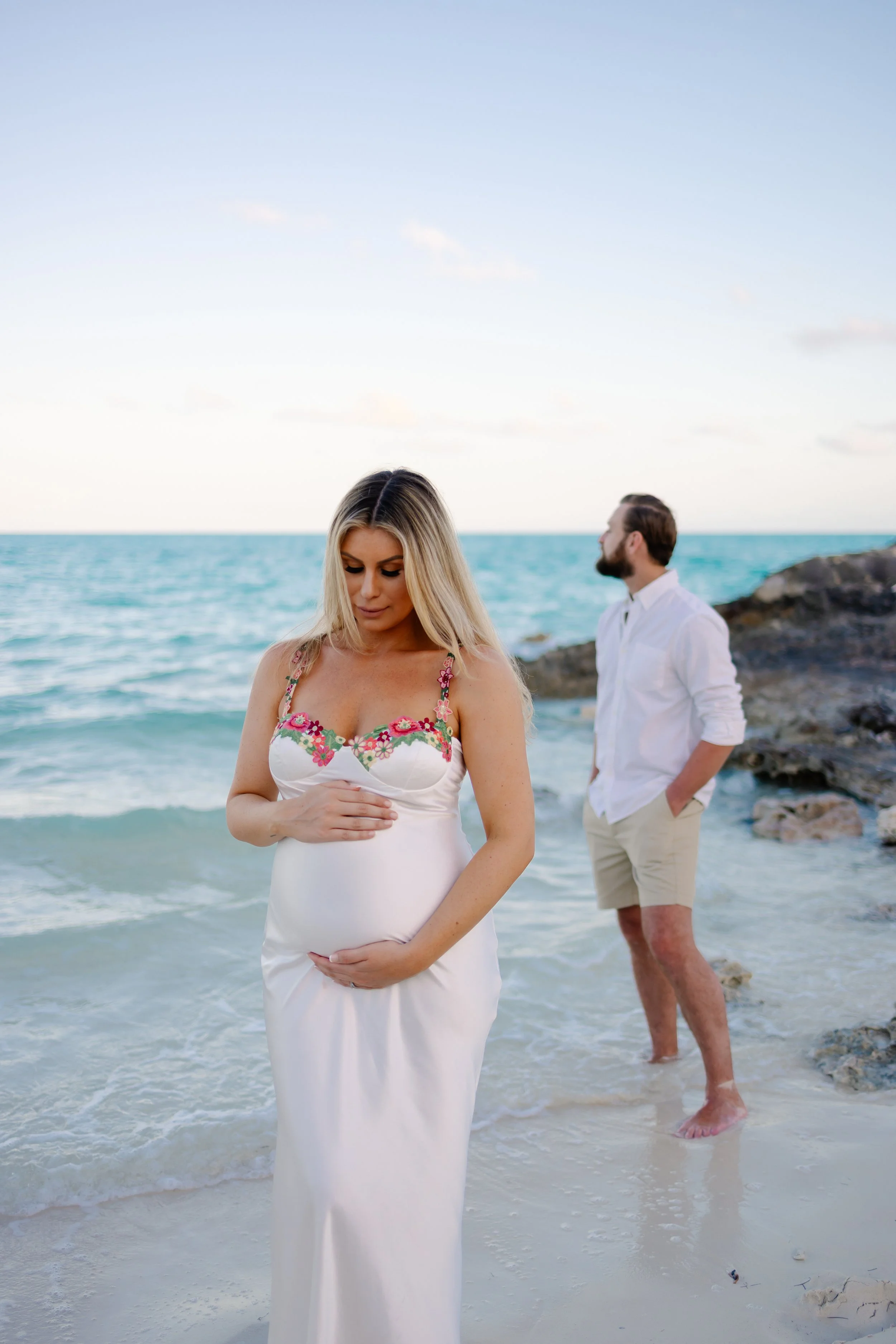 A pregnant woman in a white dress with pink floral embroidery holding her belly on a beach, with a man in light-colored shorts and a white shirt standing on rocks in the background.