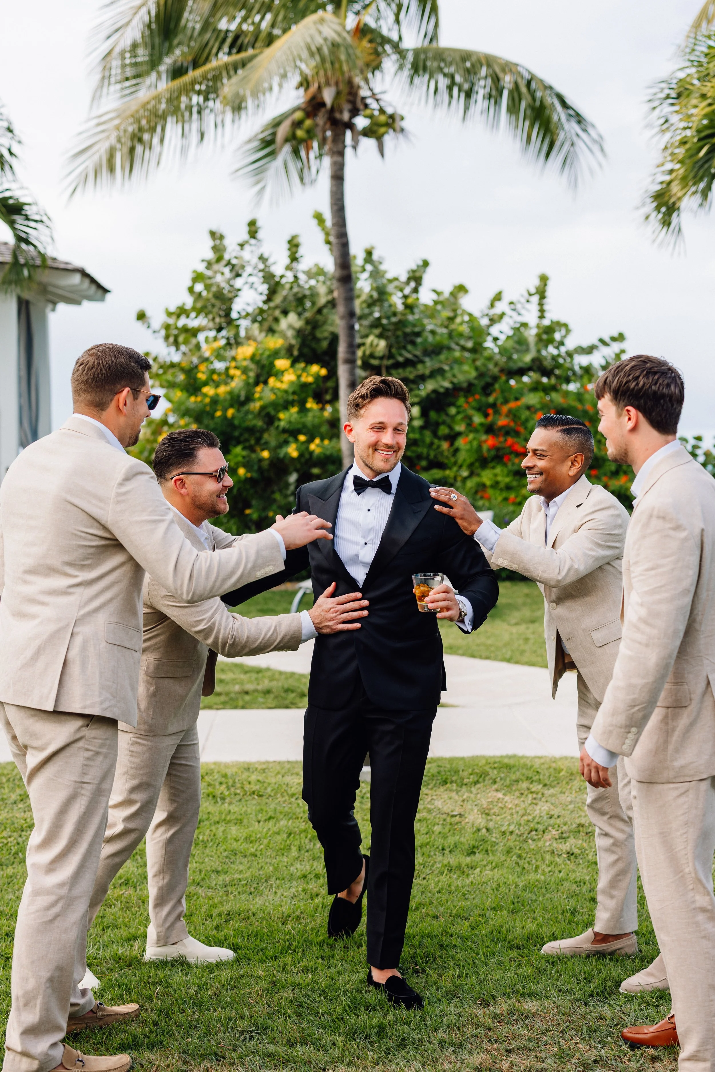 A group of six men dressed in formal suits having a celebration outdoors with a tropical background of palm trees and colorful flowering bushes.