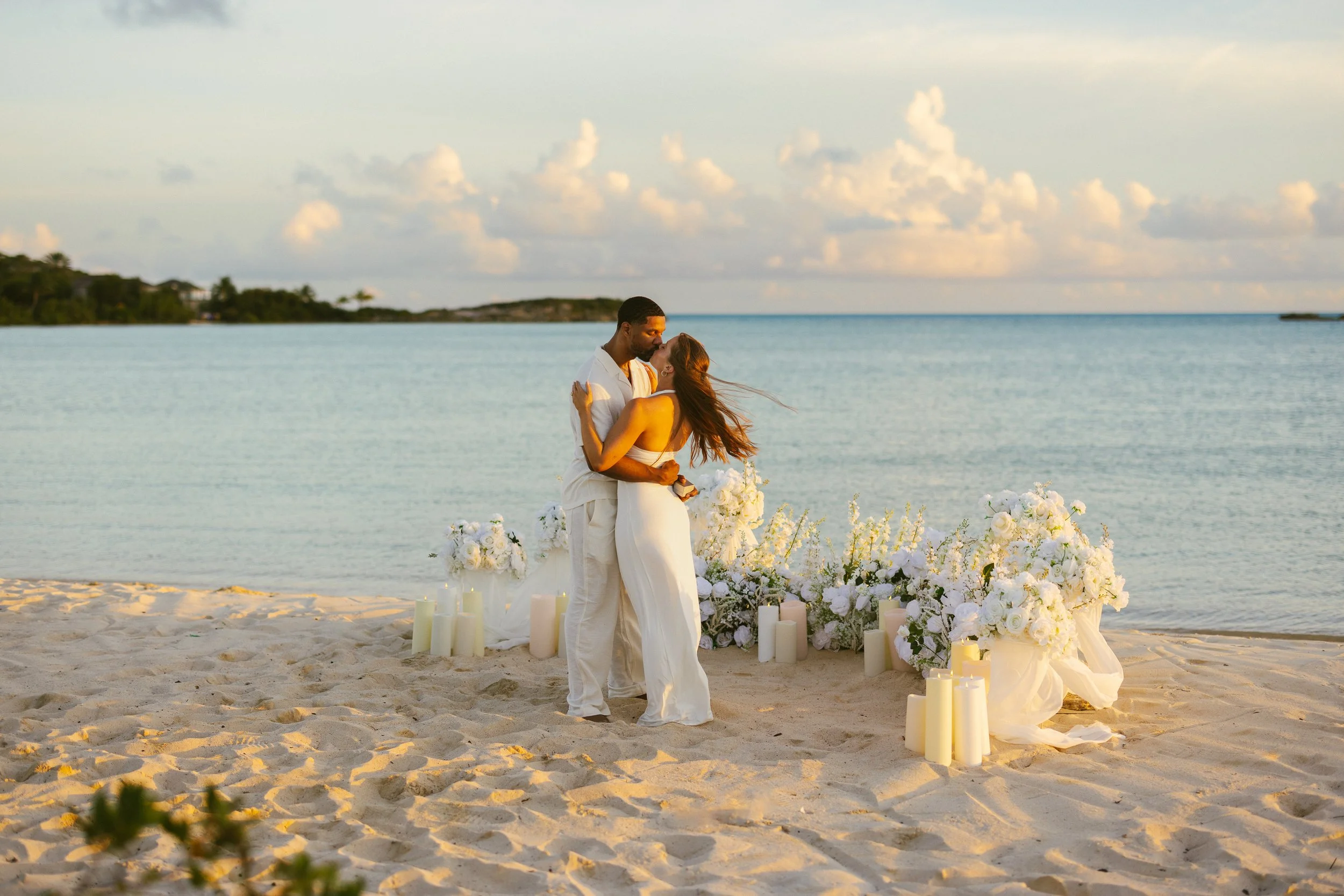 A couple dressed in white embrace on a beach during their wedding, with white floral arrangements and candles in the foreground and the ocean and a cloudy sky in the background.