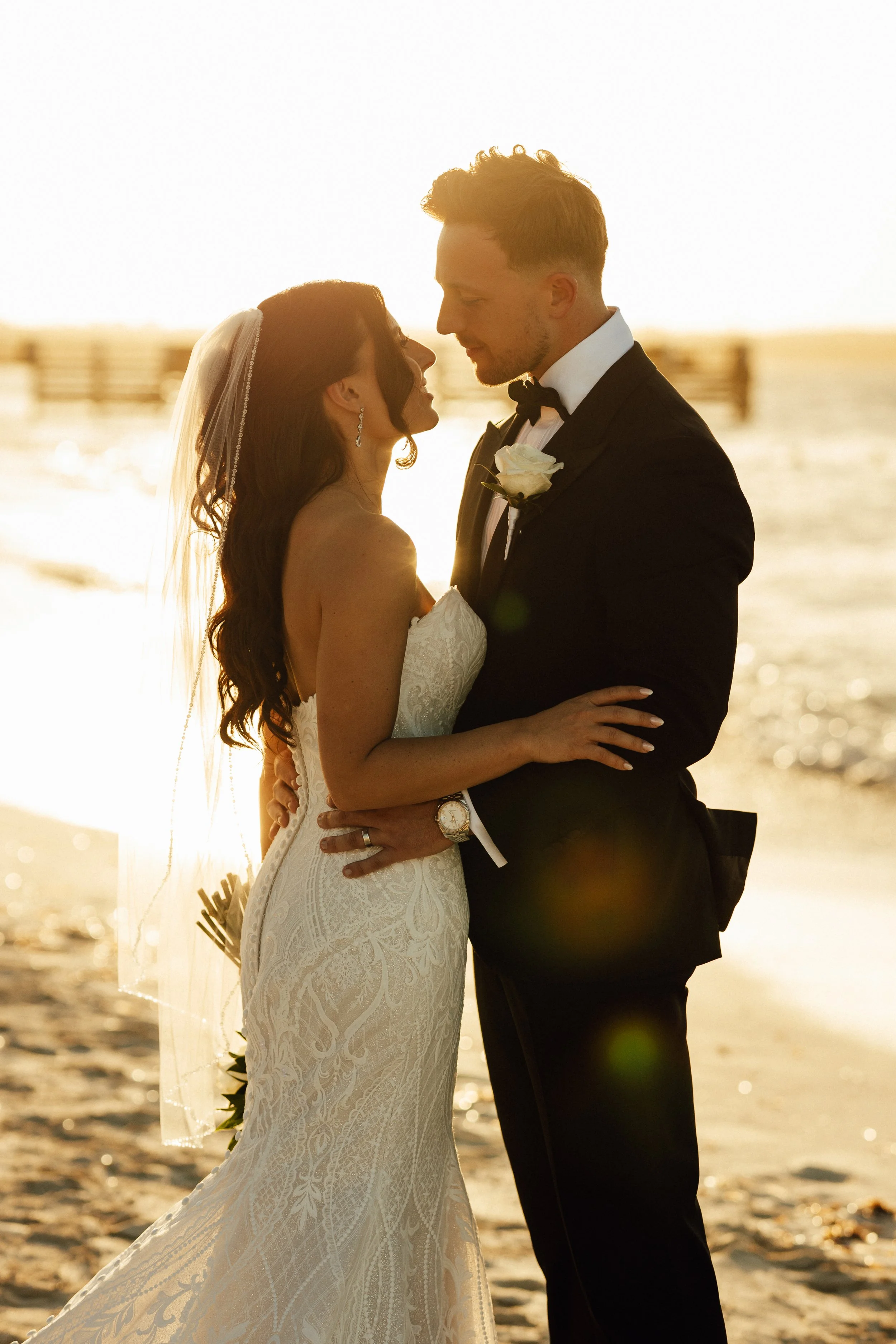 A bride and groom in wedding attire sharing a kiss on the beach during sunset with the ocean in the background.