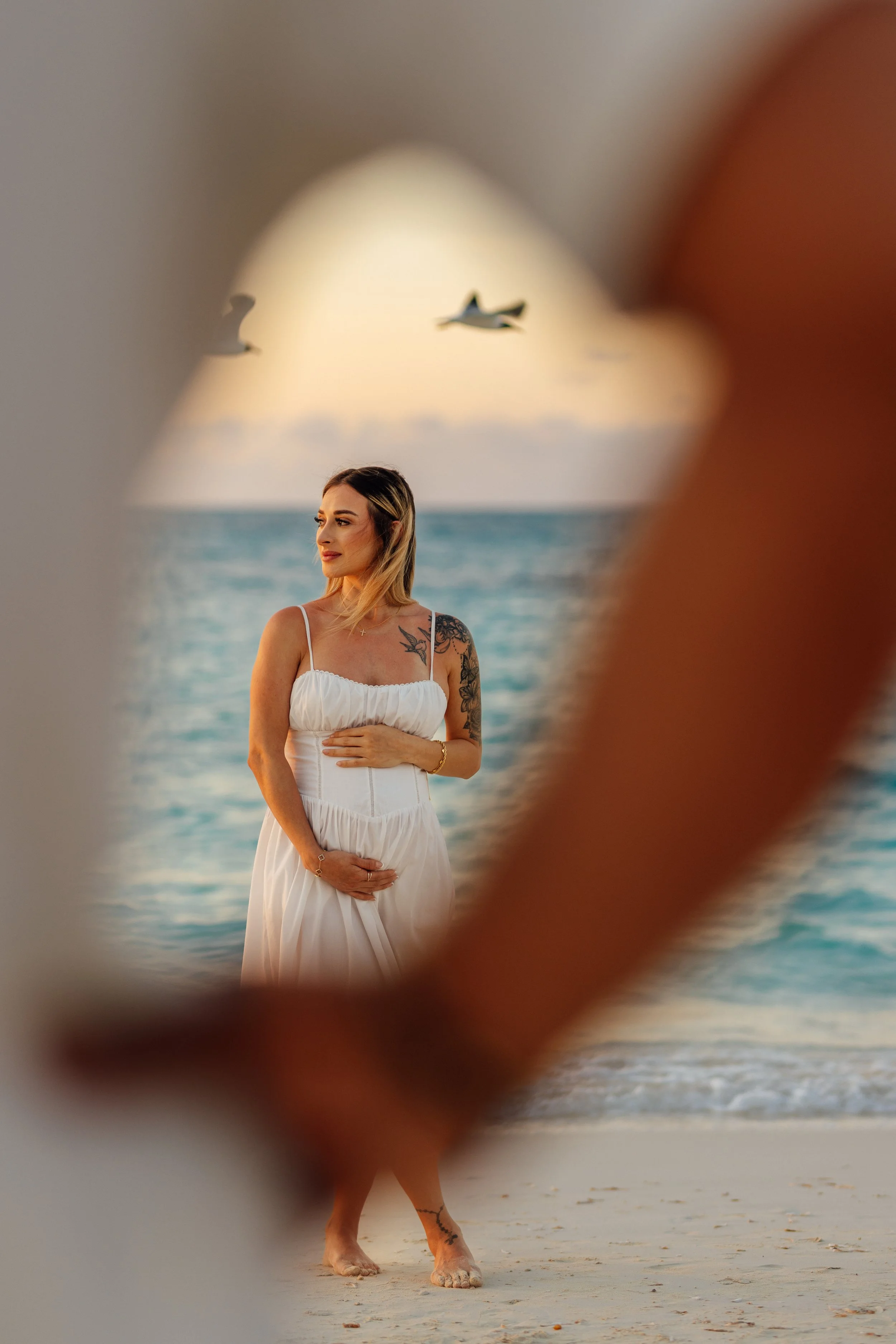 A woman standing on the beach, partially obscured by a frame, with ocean and sky in the background. The woman has long hair, tattoos on her right arm, and is wearing a white dress. Two planes are flying in the sky.