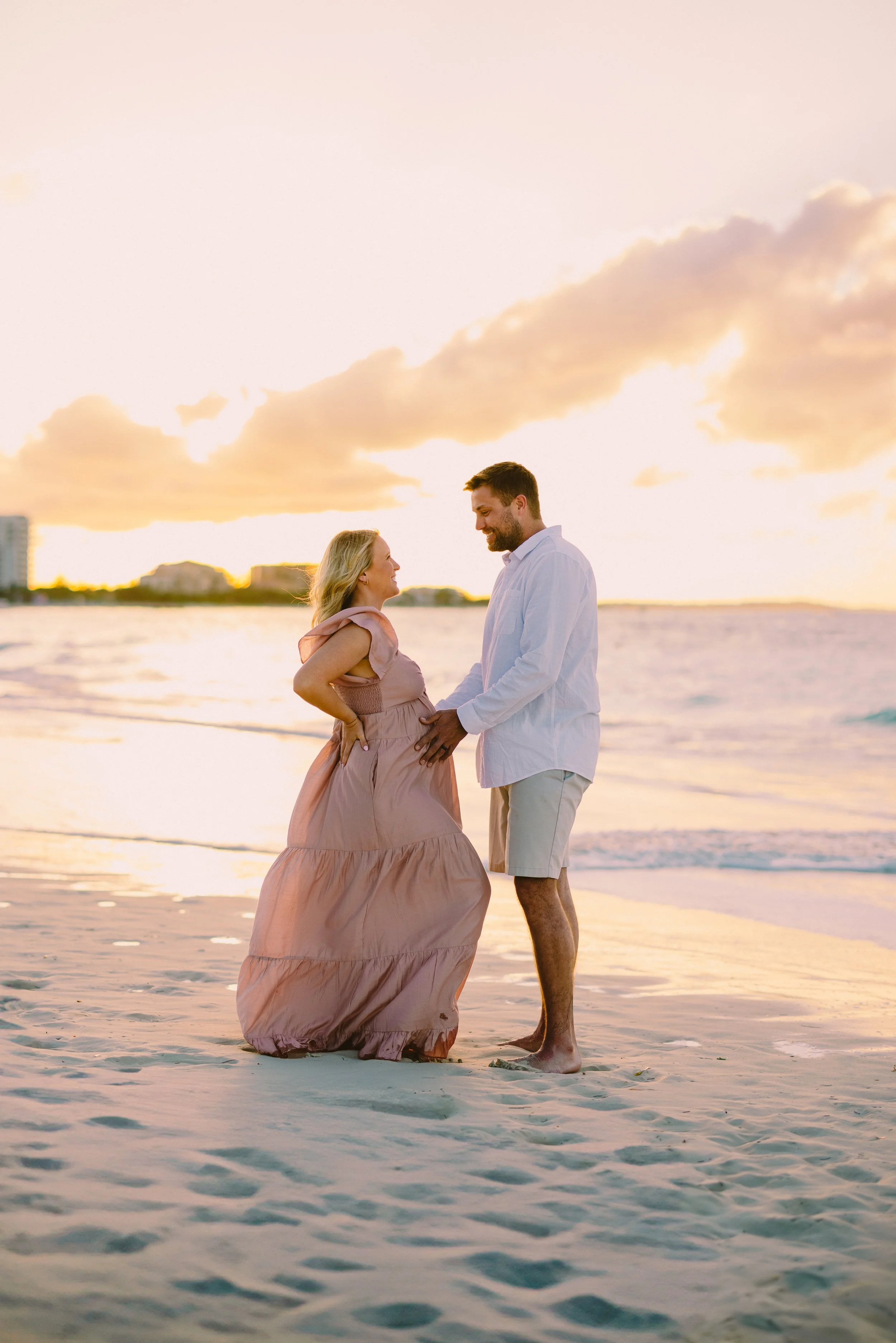 A couple standing on the beach at sunset, looking at each other and holding hands, with the woman pregnant, wearing a pink dress and the man in a white shirt and shorts.