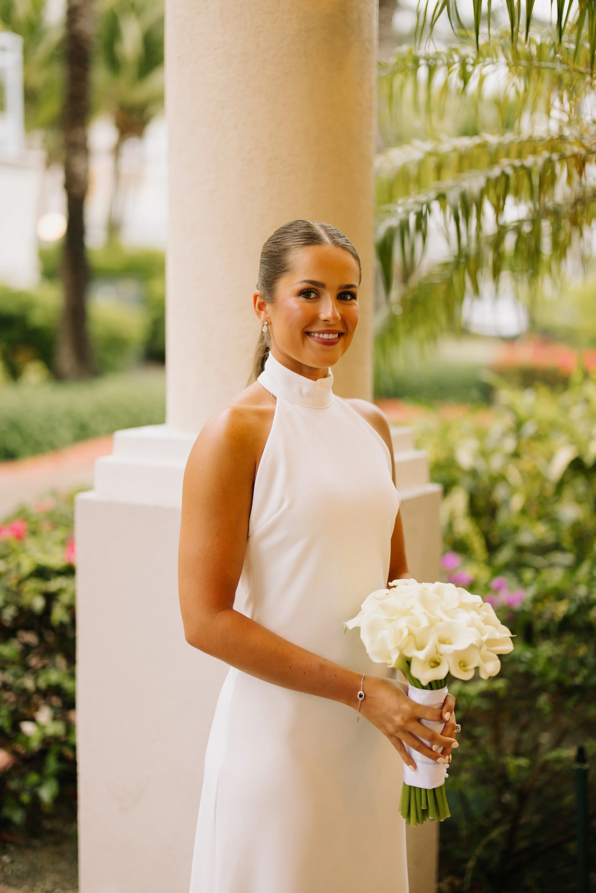 A woman in a white dress holding a bouquet of white flowers outdoors, with greenery and a column in the background.