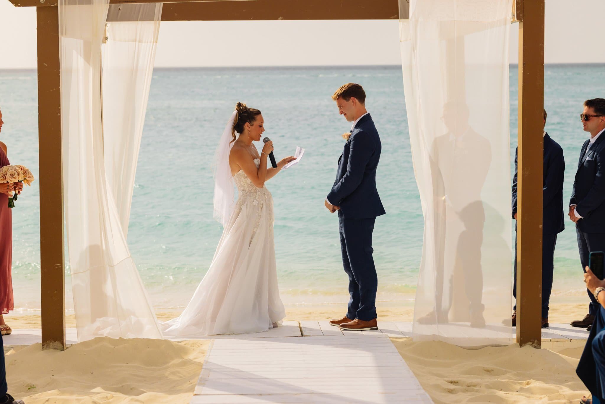 A bride and groom exchanging vows during a beach wedding, with the bride holding a microphone and reading from a paper, standing under a canopy with white drapes, ocean in the background, and several onlookers on either side.
