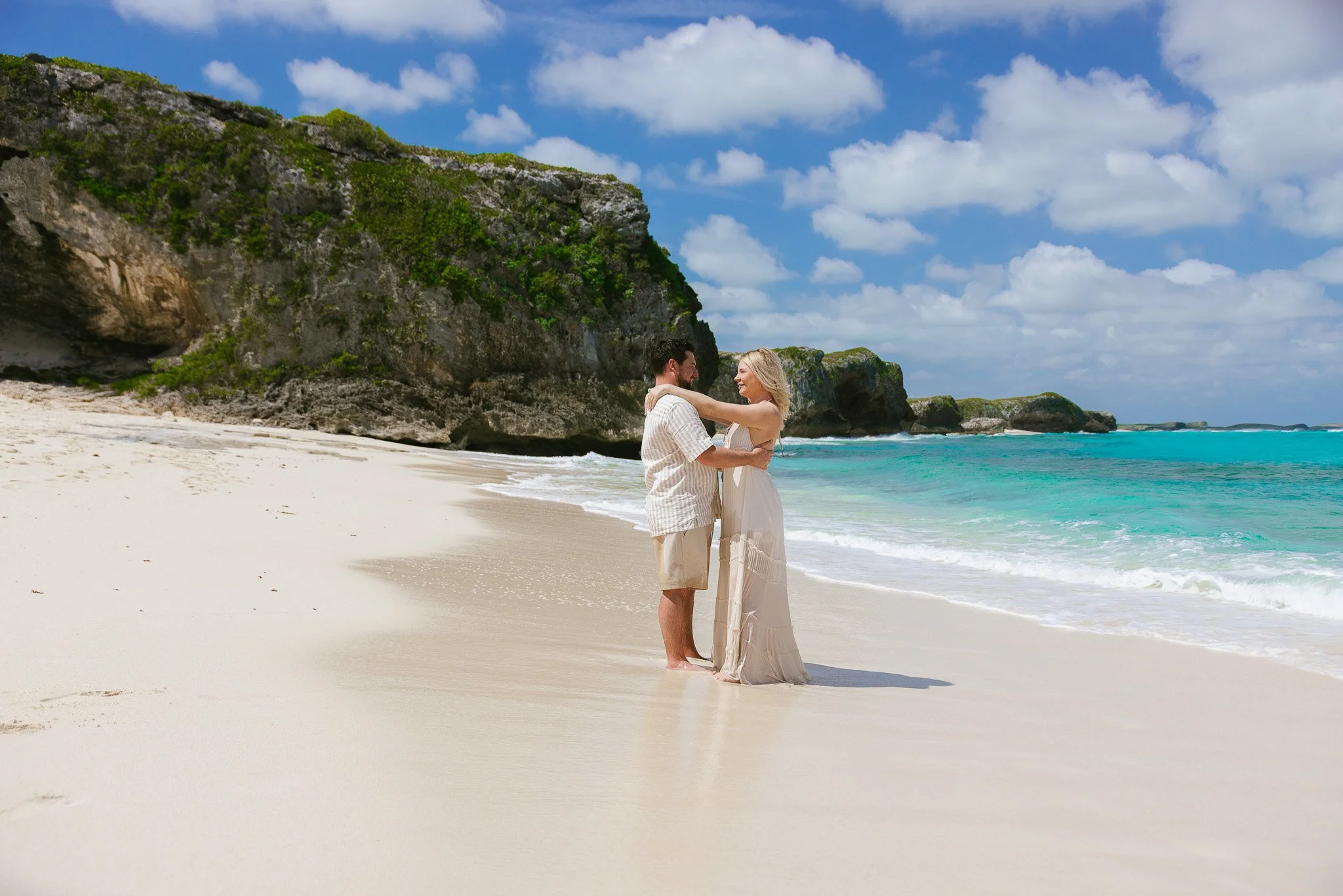 A couple stands on a sandy beach, embracing and smiling at each other. The woman wears a white dress, and the man is dressed in a light-colored shirt and shorts. The background showcases turquoise water, green cliffs, and a partly cloudy sky.