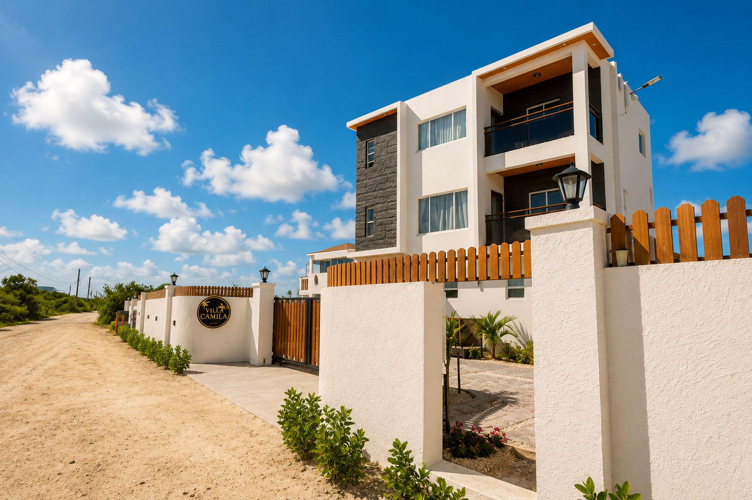 Modern white and gray multi-story building with balconies, surrounded by a white wall with wood accents and a black gate, under a clear blue sky with clouds.