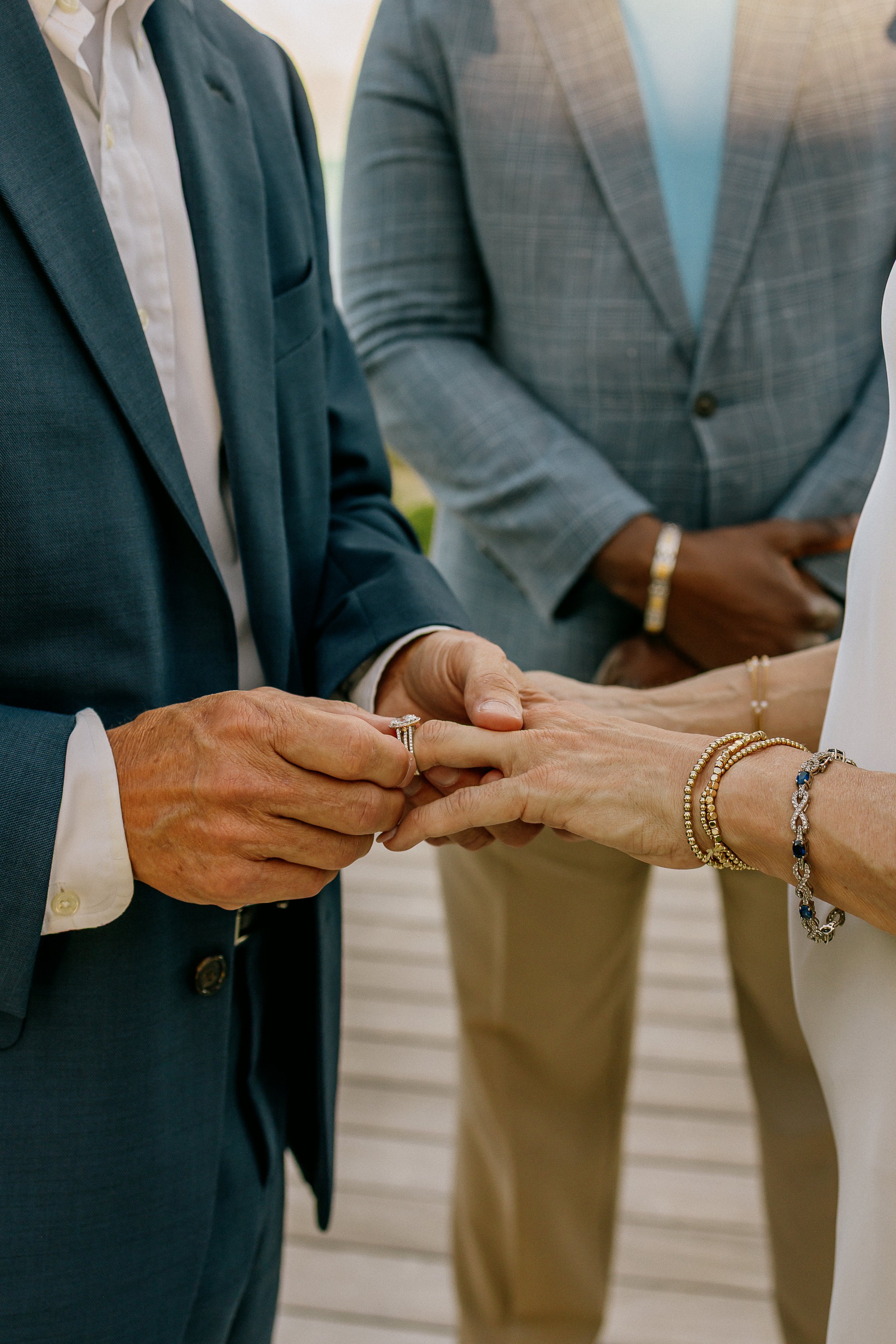 Close-up of a man placing a ring on a woman's finger during a wedding ceremony, with the couple's hands and jewelry in focus and the officiant and guests in the background.