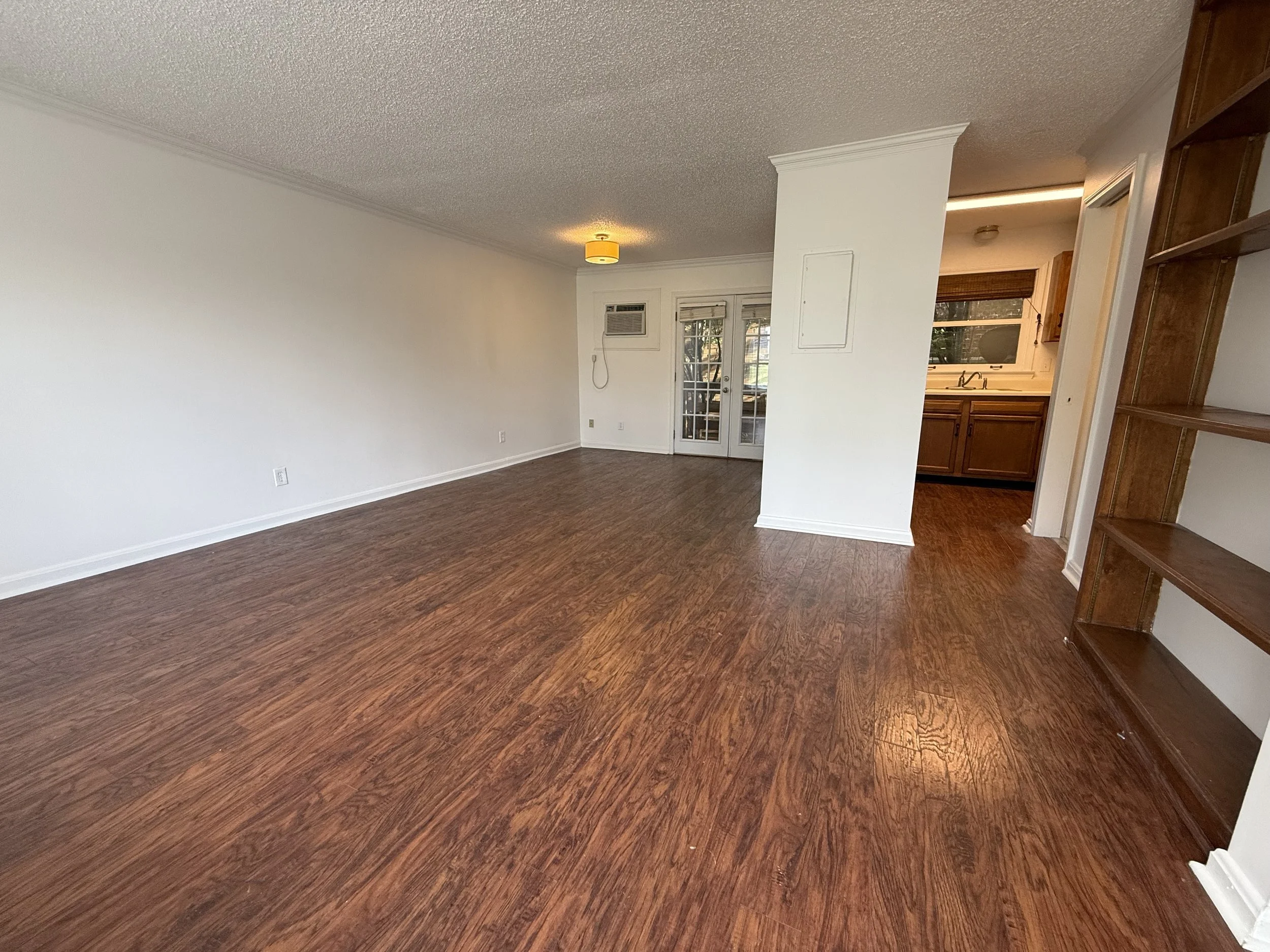 Empty living room with wood flooring, white walls, a ceiling light, a door with windows, and a kitchen area with wooden cabinets visible to the right.