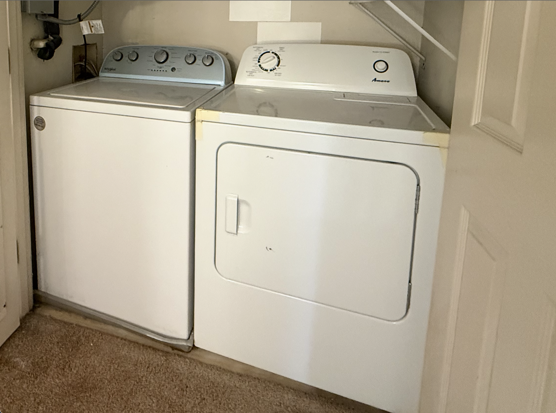 A laundry room with a top-loading washing machine on the left and a top-loading dryer on the right, both white, with beige walls and carpeted floor.