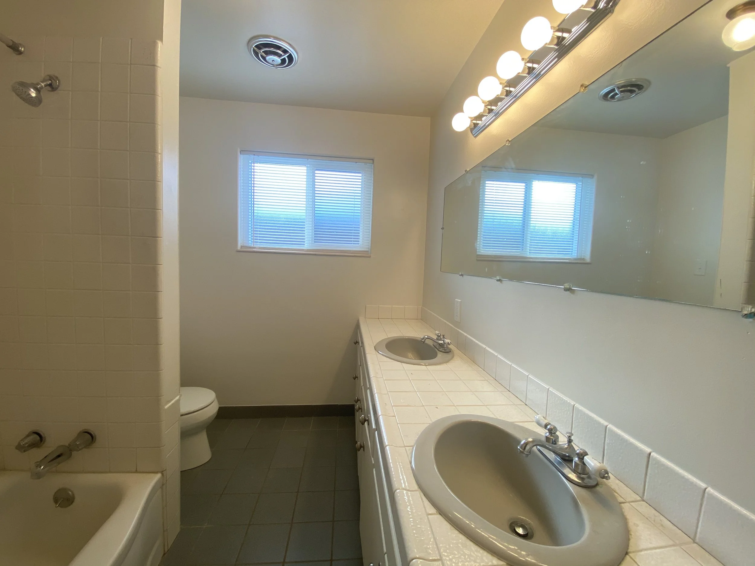 A bathroom with a double sink vanity with a mirror above it, two windows with blinds, a toilet, and a shower with a wall of square tiles. The room has a light fixture with multiple round bulbs overhead.