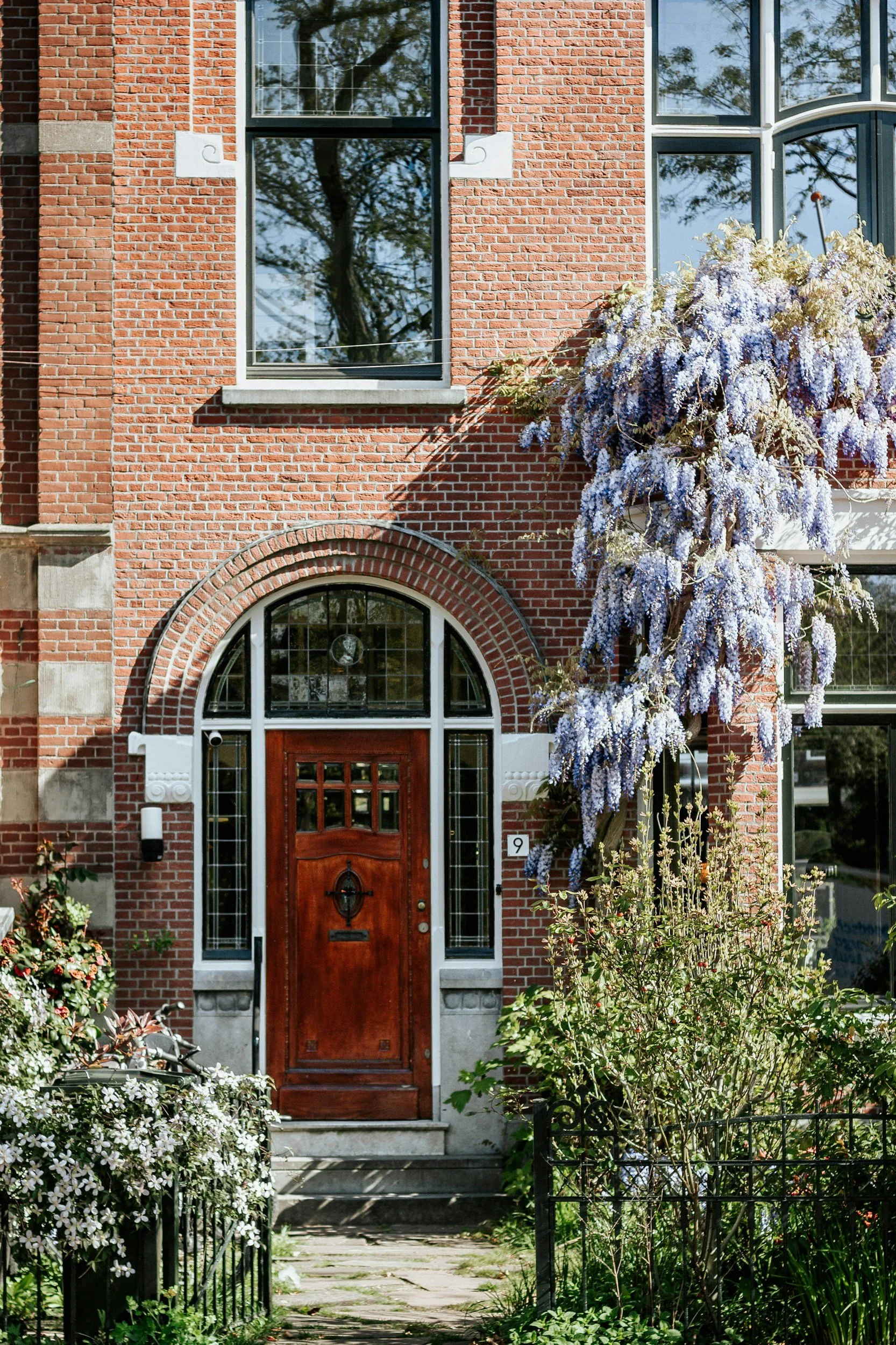 Brick house with an arched wooden front door, large windows, and a lush garden with flowering bushes and trees.