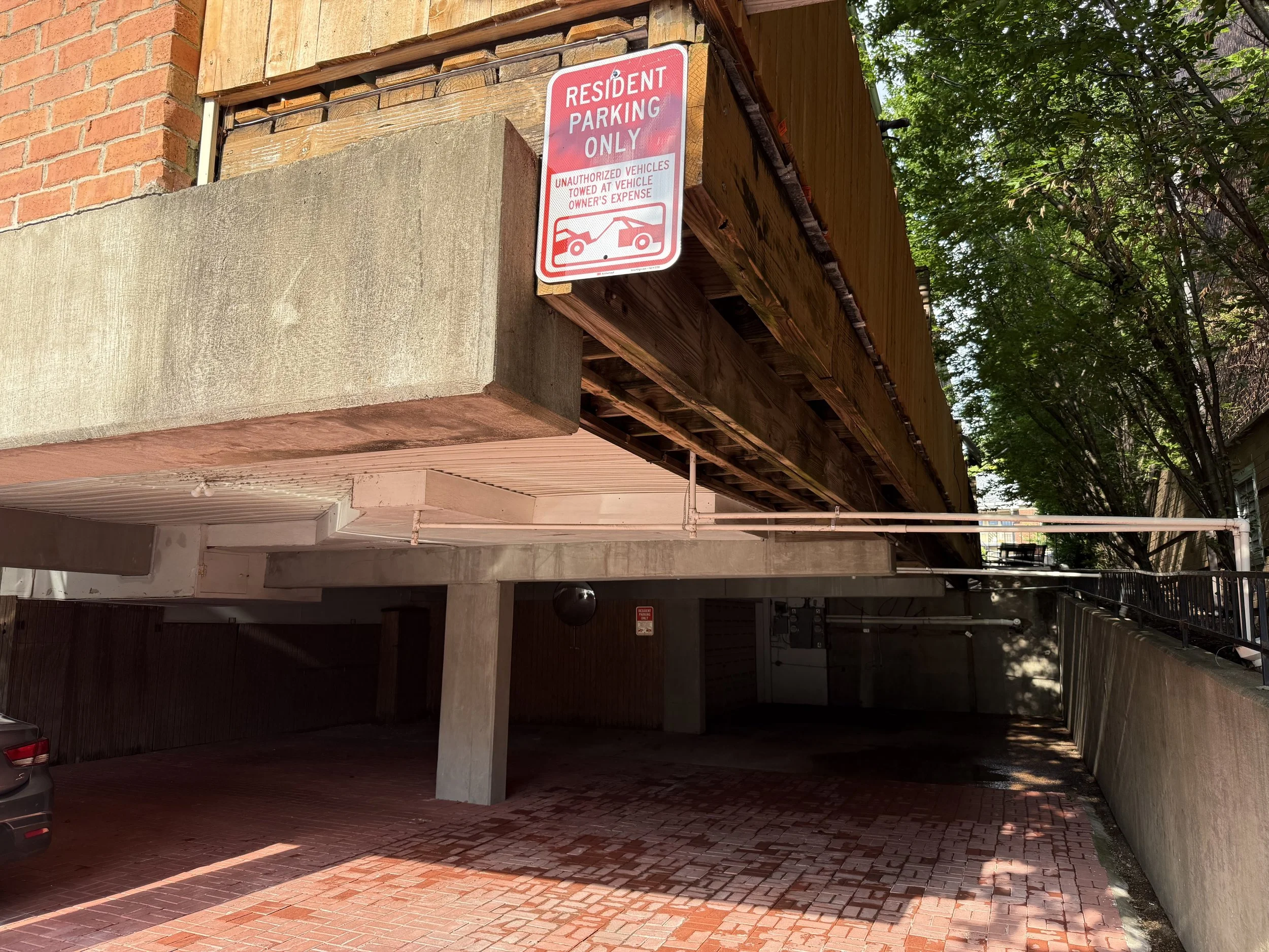 Parking garage with a sign that says 'Resident Parking Only' and warns unauthorized vehicles will be towed at vehicle owner's expense. The garage has brick and concrete walls, and is partially shaded by trees.