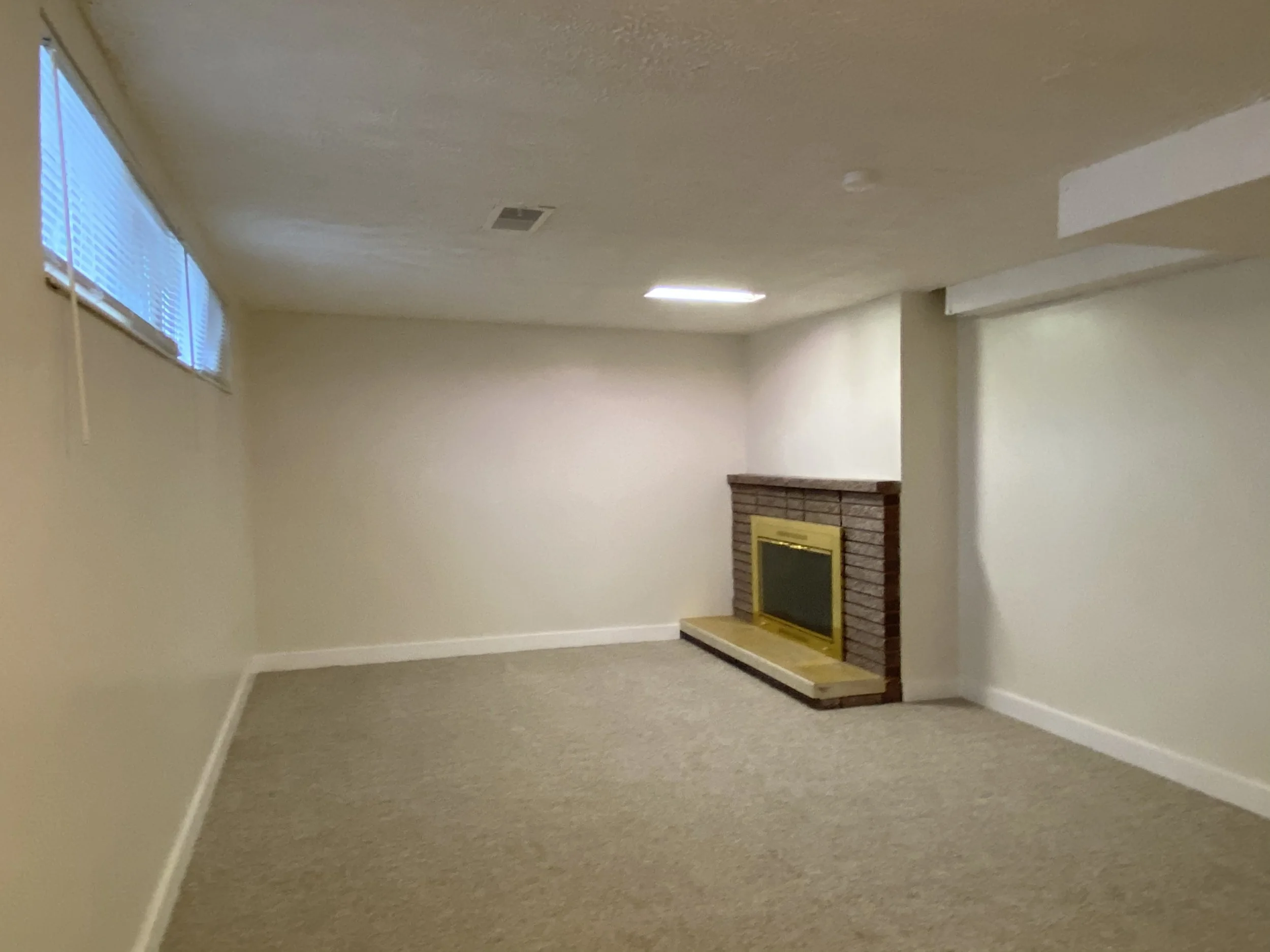 Empty room with a brick fireplace, corner windows with blinds, beige walls, and carpeted floor.