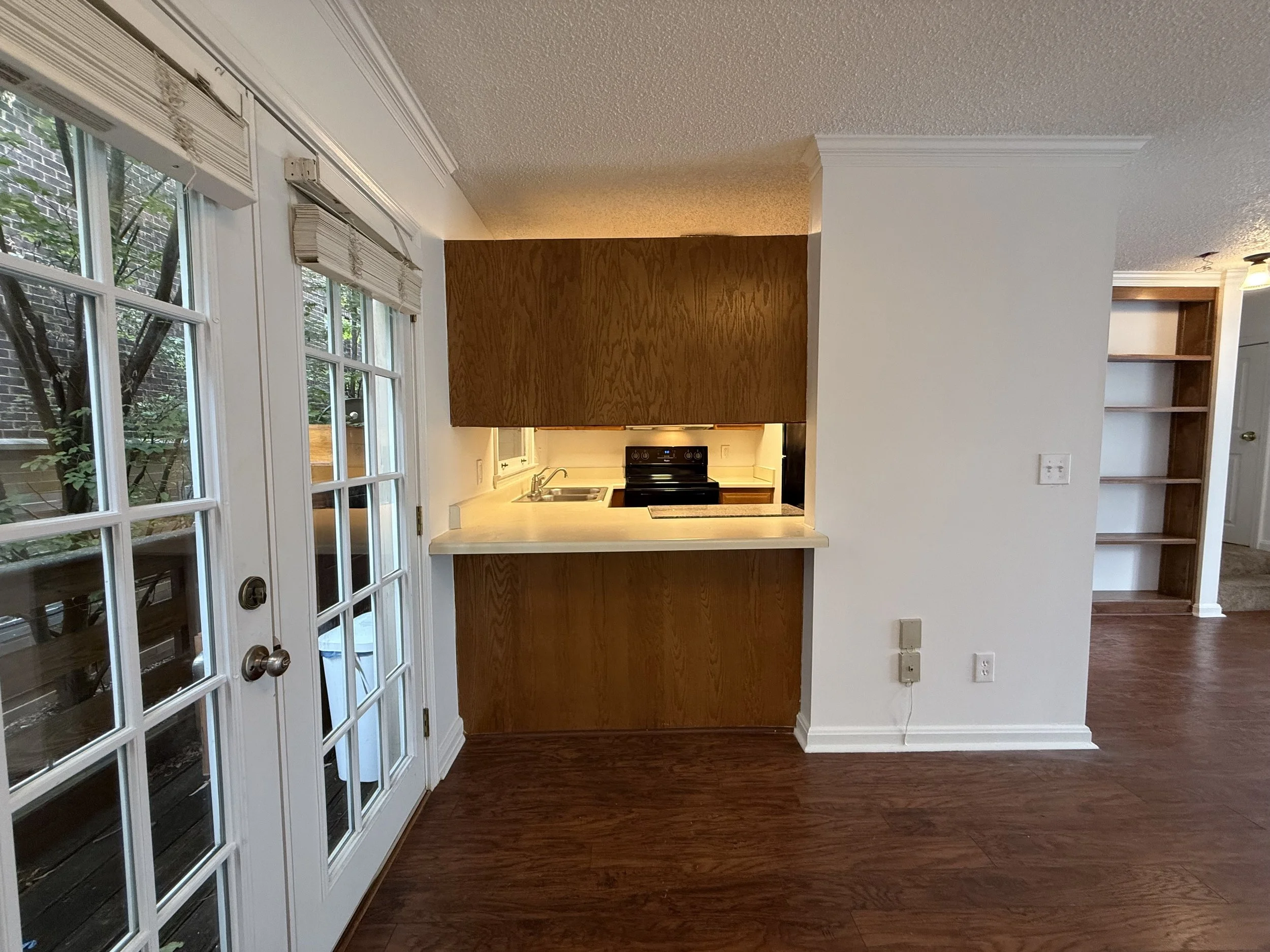 Interior view of a kitchen with wooden cabinets, a stove, and a white countertop, seen from a room with brown wood floors and a glass-paneled door leading outside.