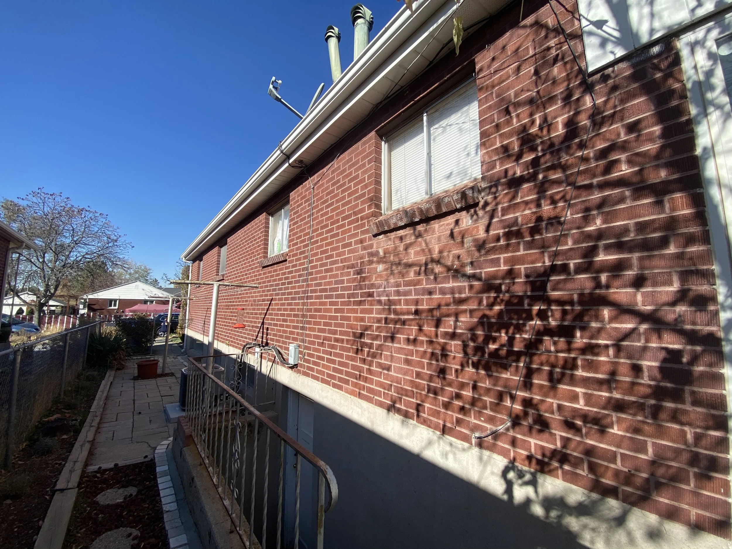 The side of a brick house with a window, an outdoor air conditioning unit below the window, and a shadow of tree branches on the brick wall. There is a small patio area with pavers, a fence, and some potted plants. The sky is clear and blue.