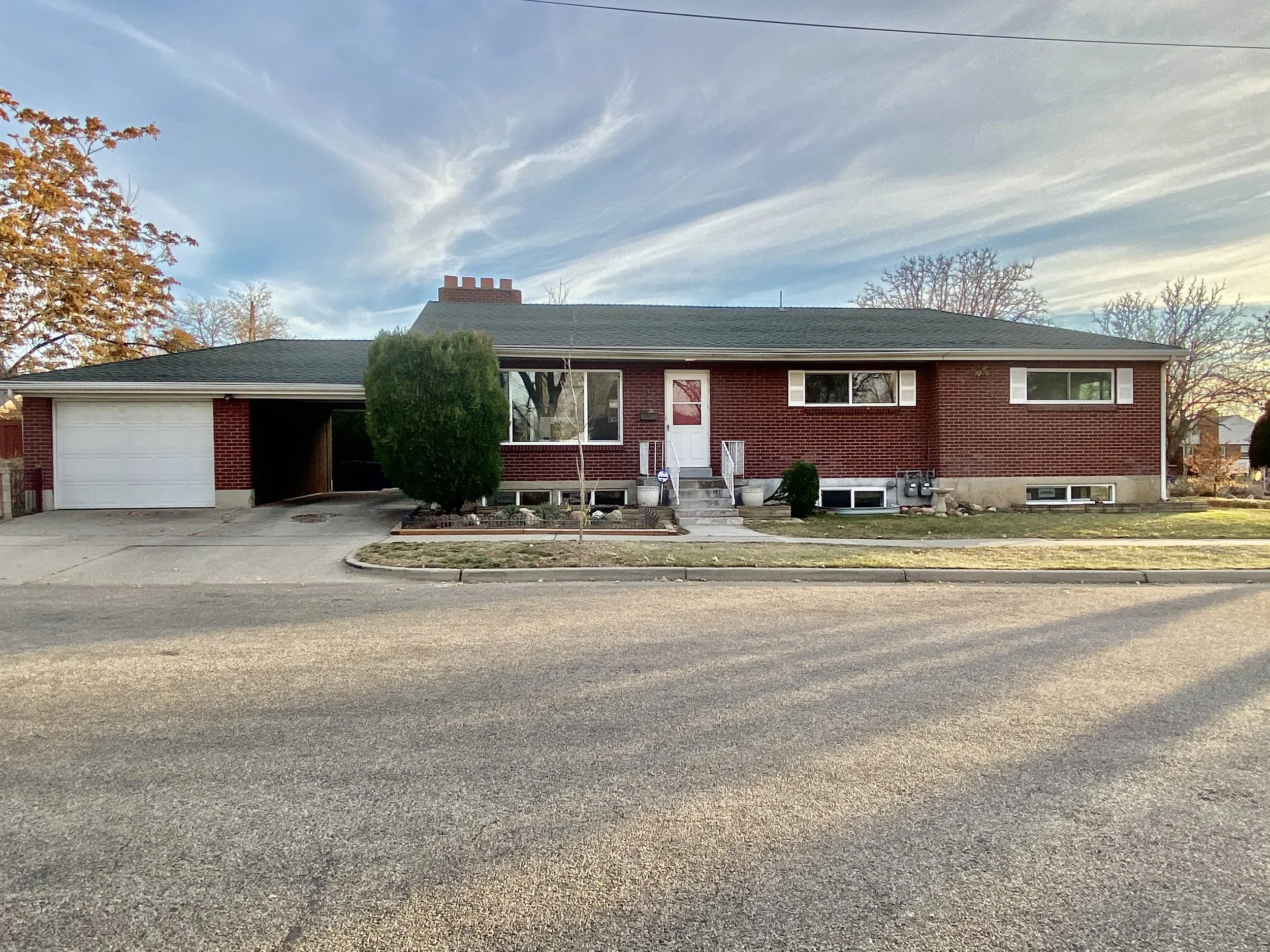 A brick house with a garage on the left side, a front door with stairs, and multiple windows. There are trees with autumn leaves on the left and right sides, and the sky is partially cloudy with visible clouds and a slight sunset glow.