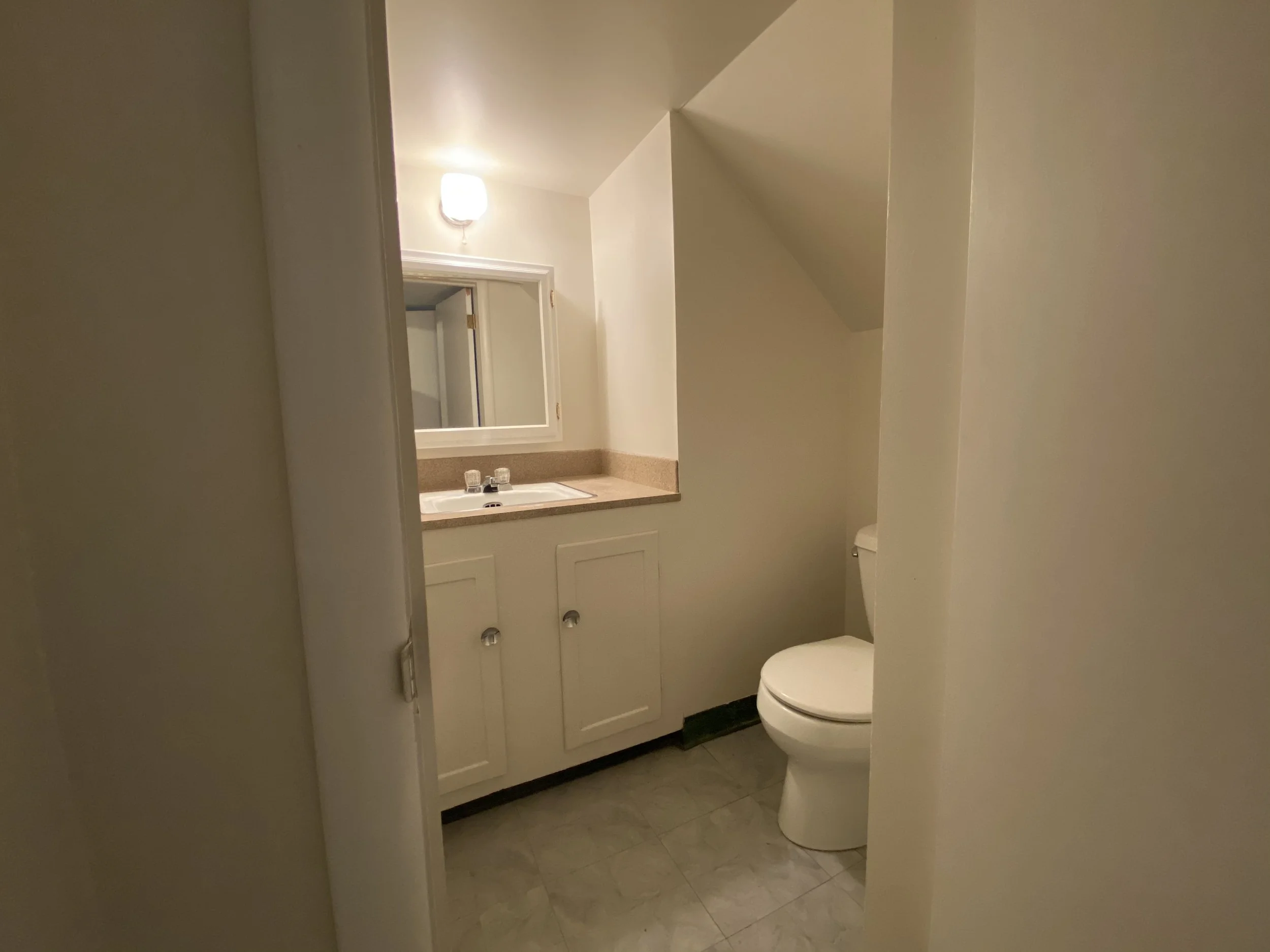 A small bathroom with a white toilet, a beige countertop with a sink, a mirror above the sink, and a wall-mounted light fixture, with an angular ceiling and a door frame partially visible.