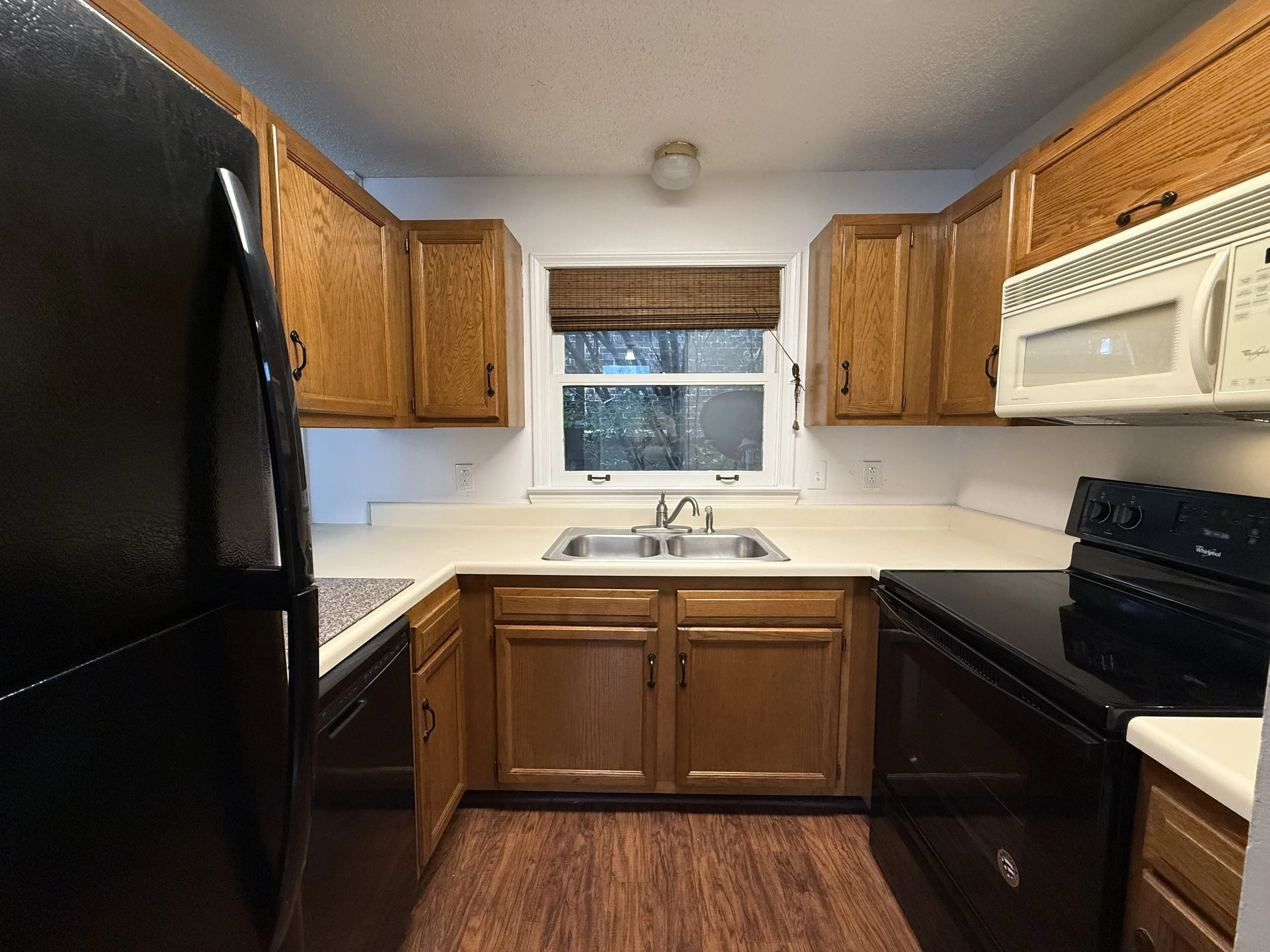 A kitchen with wooden cabinets, a white microwave, black appliances, a window with a brown shade, and a stainless steel sink.