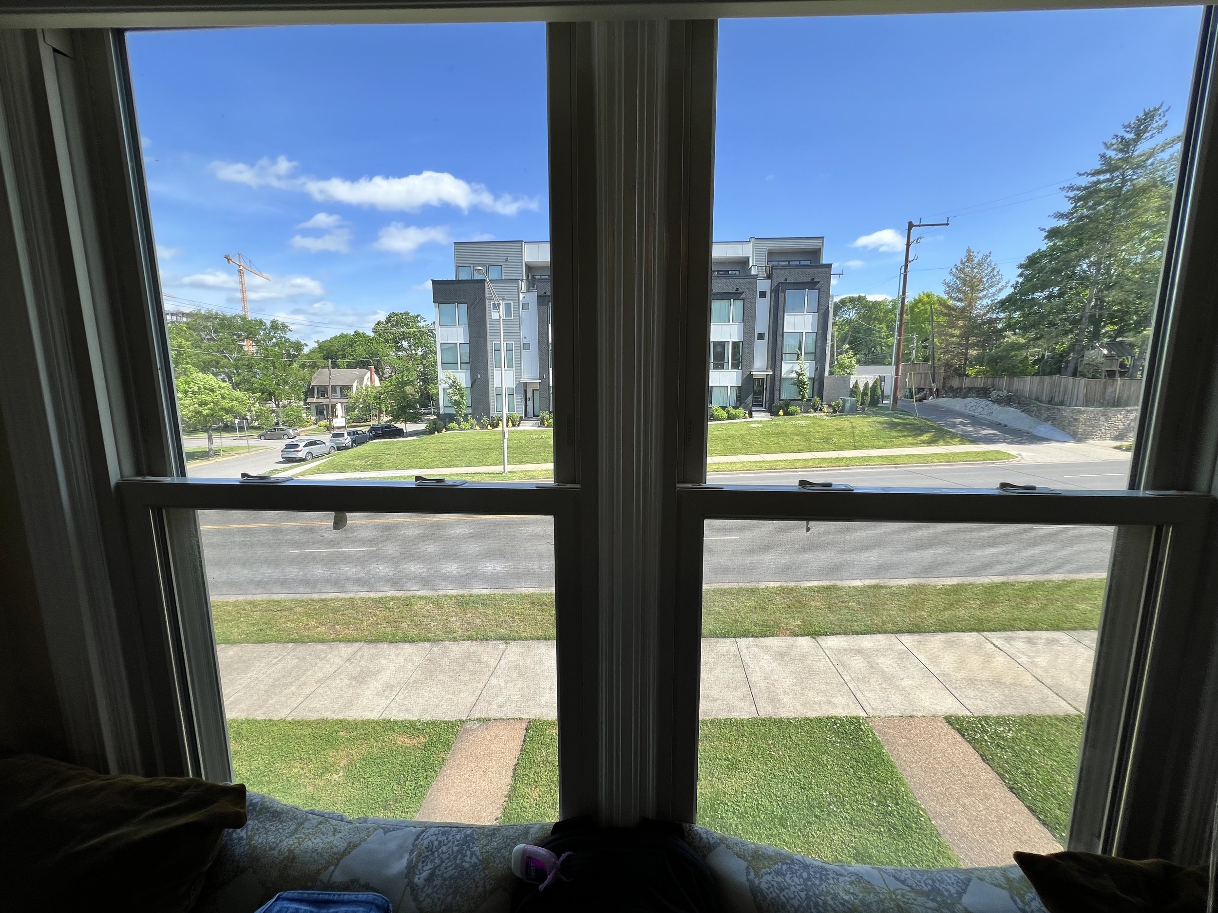 View from inside a building looking out a window at a residential street with parked cars, green lawns, trees, modern apartment buildings, and utility poles under a bright blue sky with some clouds.
