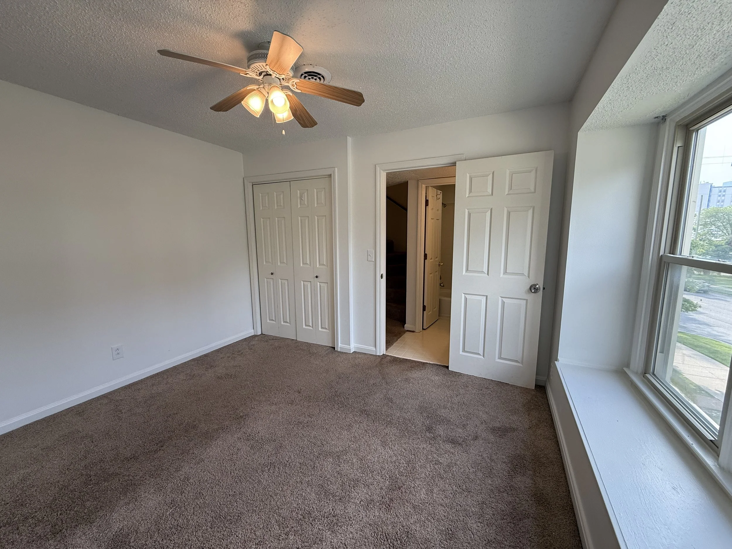 Empty bedroom with beige carpet, white walls, a ceiling fan with lights, closet doors, an open door leading to another room, and large windows showing trees and a building outside.