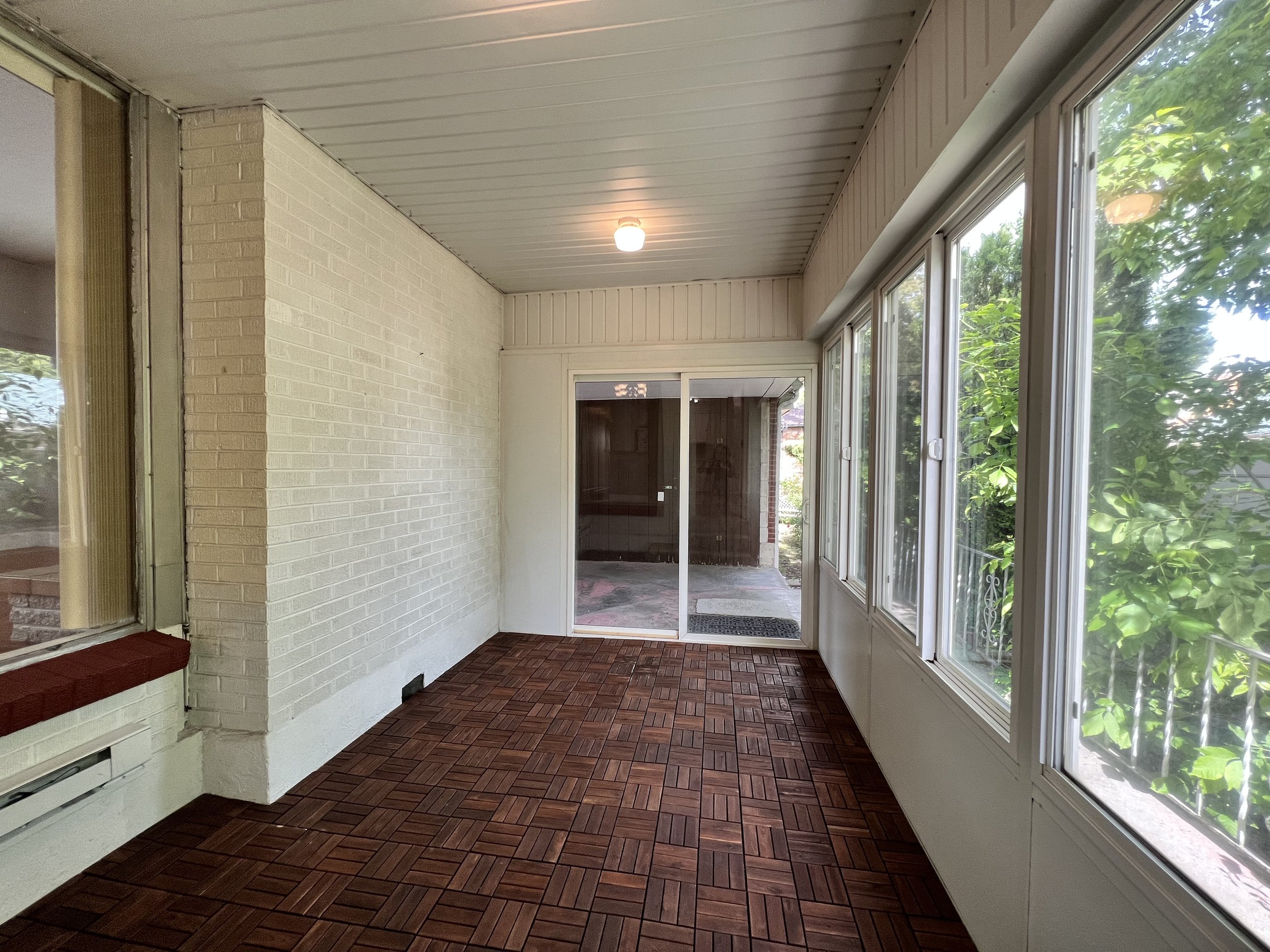 A porch area with white brick walls, glass sliding door, and large windows showing green trees outside. The ceiling has white paneling and a ceiling light, and the floor is covered with red-brown parquet tiles.
