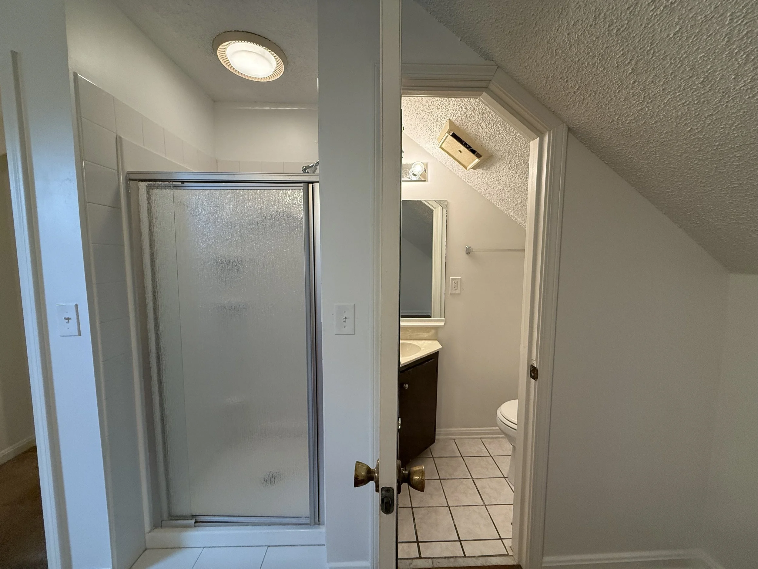 Bathroom with a frosted glass shower door, white tile flooring, a ceiling light, and a sloped ceiling with a vent. Part of a mirror and a bathroom vanity are visible through the open door.