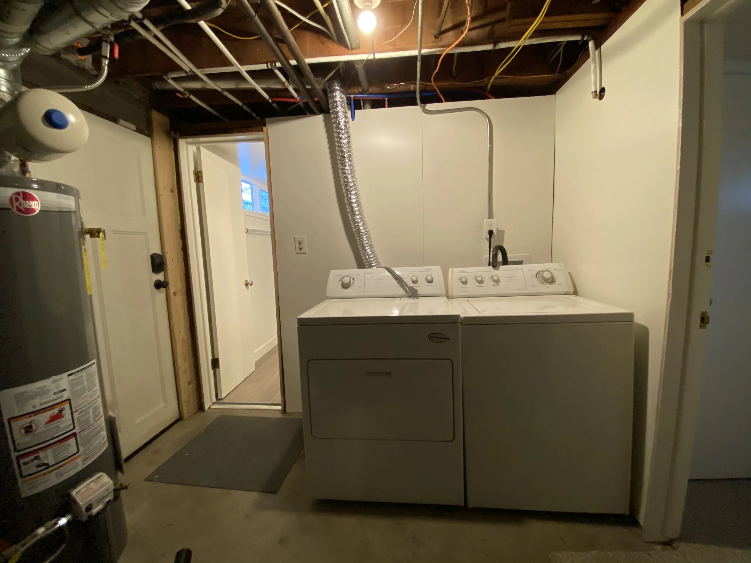 A laundry area with a white washing machine and dryer, an electrical outlet, basement ceiling exposed with pipes and wires, a wall-mounted vent duct, a water heater, and a door leading outside.