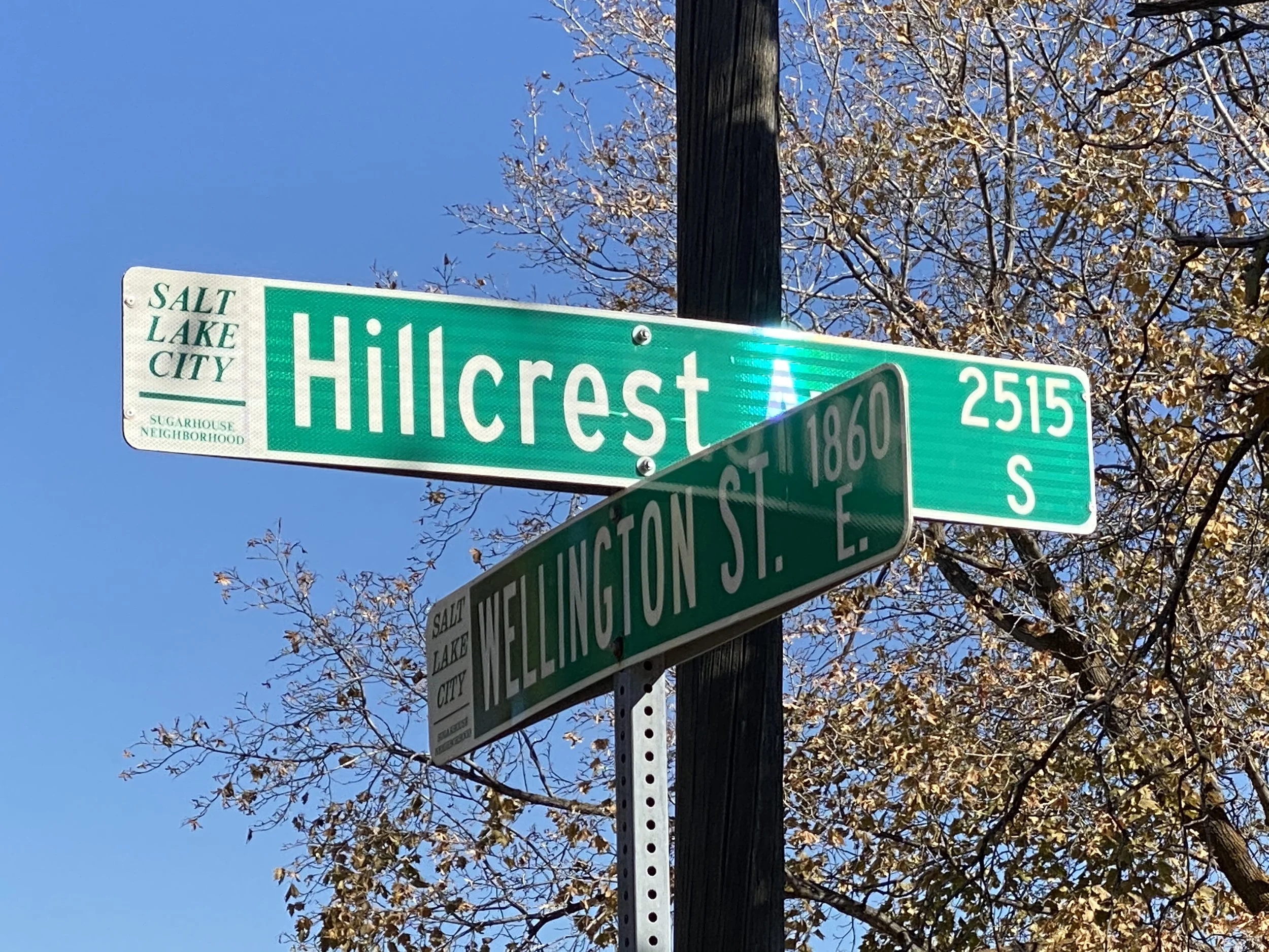Street signs at the intersection of Hillcrest Road and Wellington Street in Salt Lake City, Utah, with a bright blue sky and autumn trees in the background.