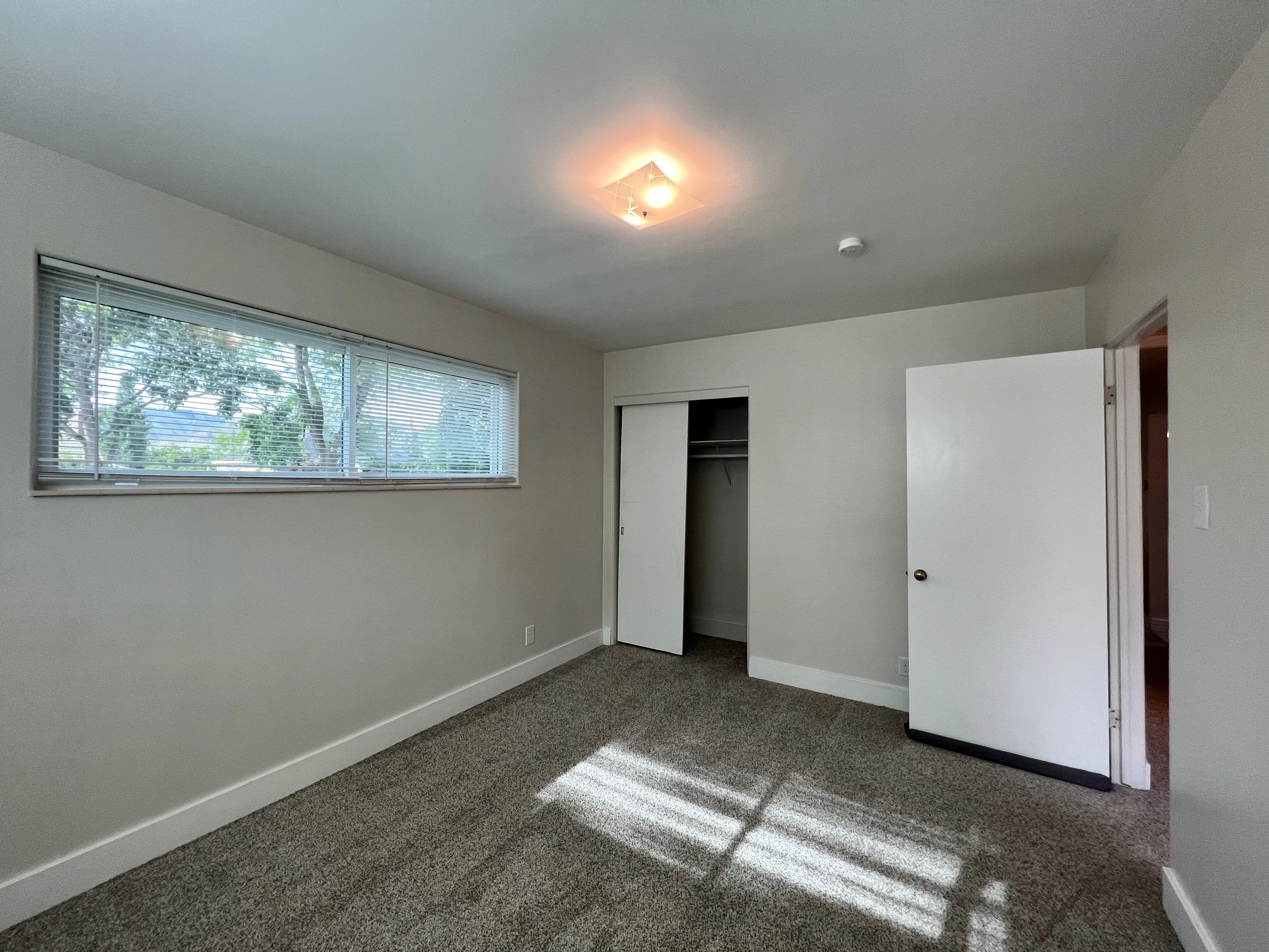 Empty room with a large window with blinds, white walls, a closet with an open door, and carpeted floor. Sunlight creates shadows on the floor.