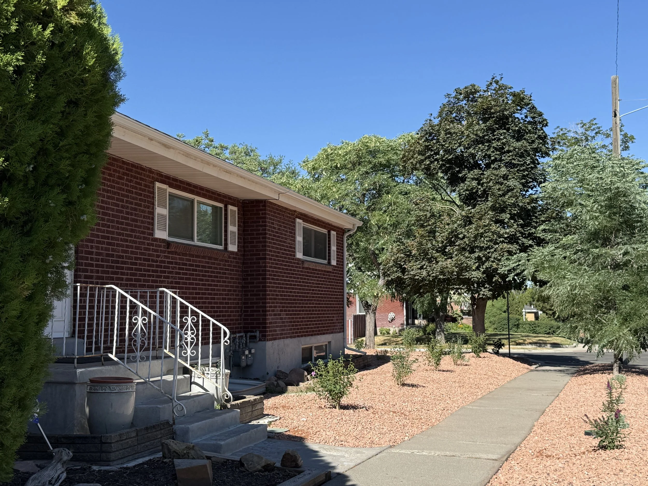 A red brick house with white window shutters, a white staircase with decorative railing, and a small landscaped yard with trees and bushes, under a clear blue sky.