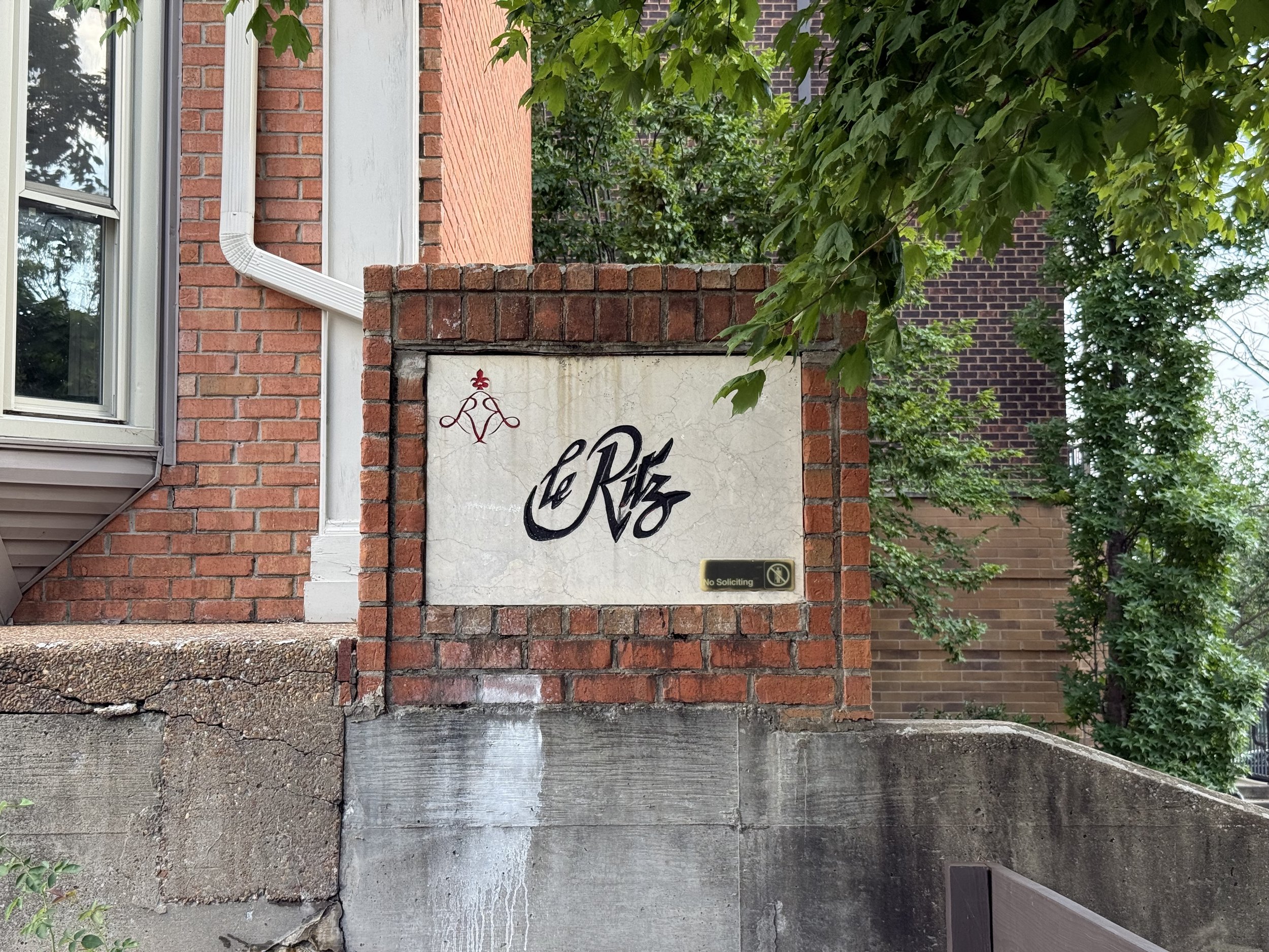 The image shows part of a brick building, including a window with white trim, a white drainpipe, and a section of a sign with decorative writing that says "le Ritz". There is a small sign below that reads "No Soliciting". Green leaves from a tree par